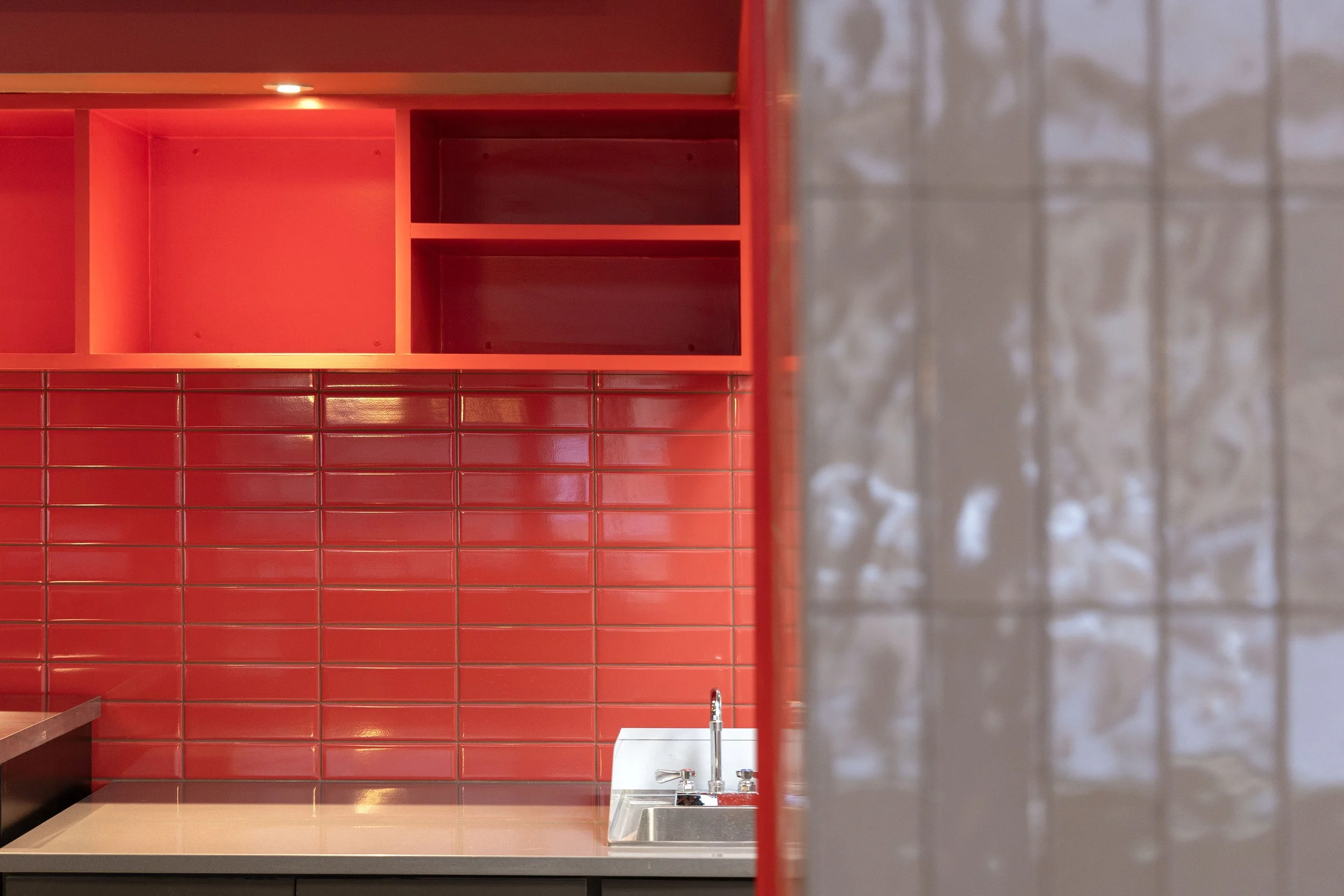 Part of a kitchen with red tiled wall, red open shelves, a white countertop, and a stainless steel sink with a faucet.