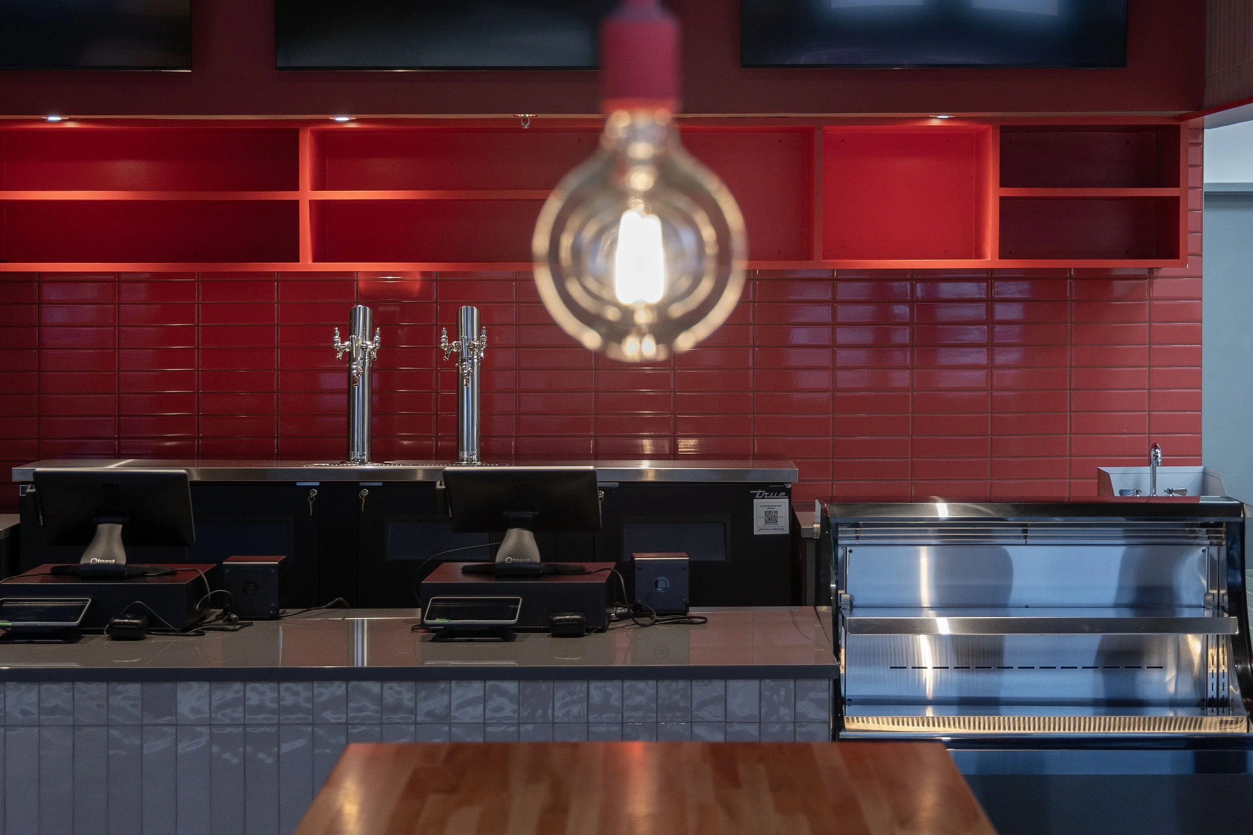 A modern cafe or bar counter with red tiled wall backdrop, two beer taps, two computer monitors, and a stainless steel food display case, illuminated by a hanging light bulb.