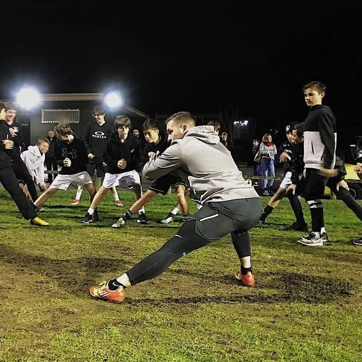 A group of kids participating in a football practice or drill at night, with a coach demonstrating a stretching or warm-up move on a grassy field.