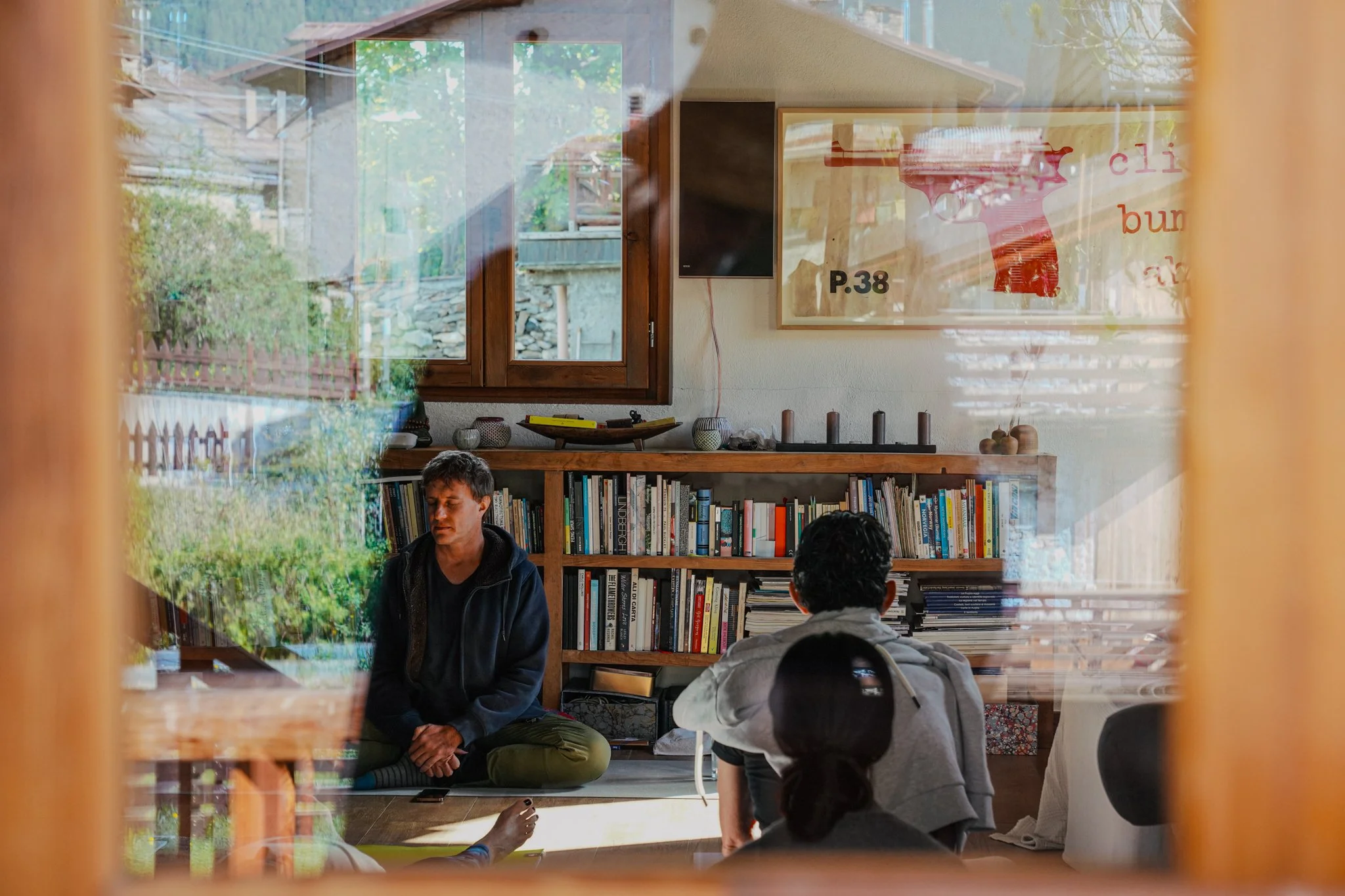 Interior of a cozy room with a person meditating, bookshelves filled with books, artwork on the wall, and sunlight reflecting on the window.