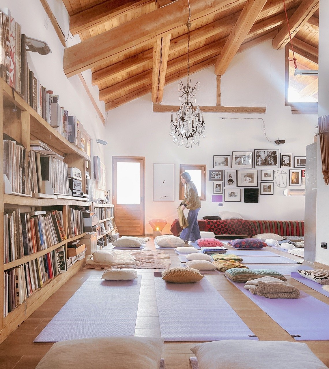 A rustic room with wooden beams, yoga mats, and cushions on the floor. Shelves filled with books line the left wall, and framed photos decorate the back wall. A woman arranges items near the back, and a chandelier hangs from the ceiling.