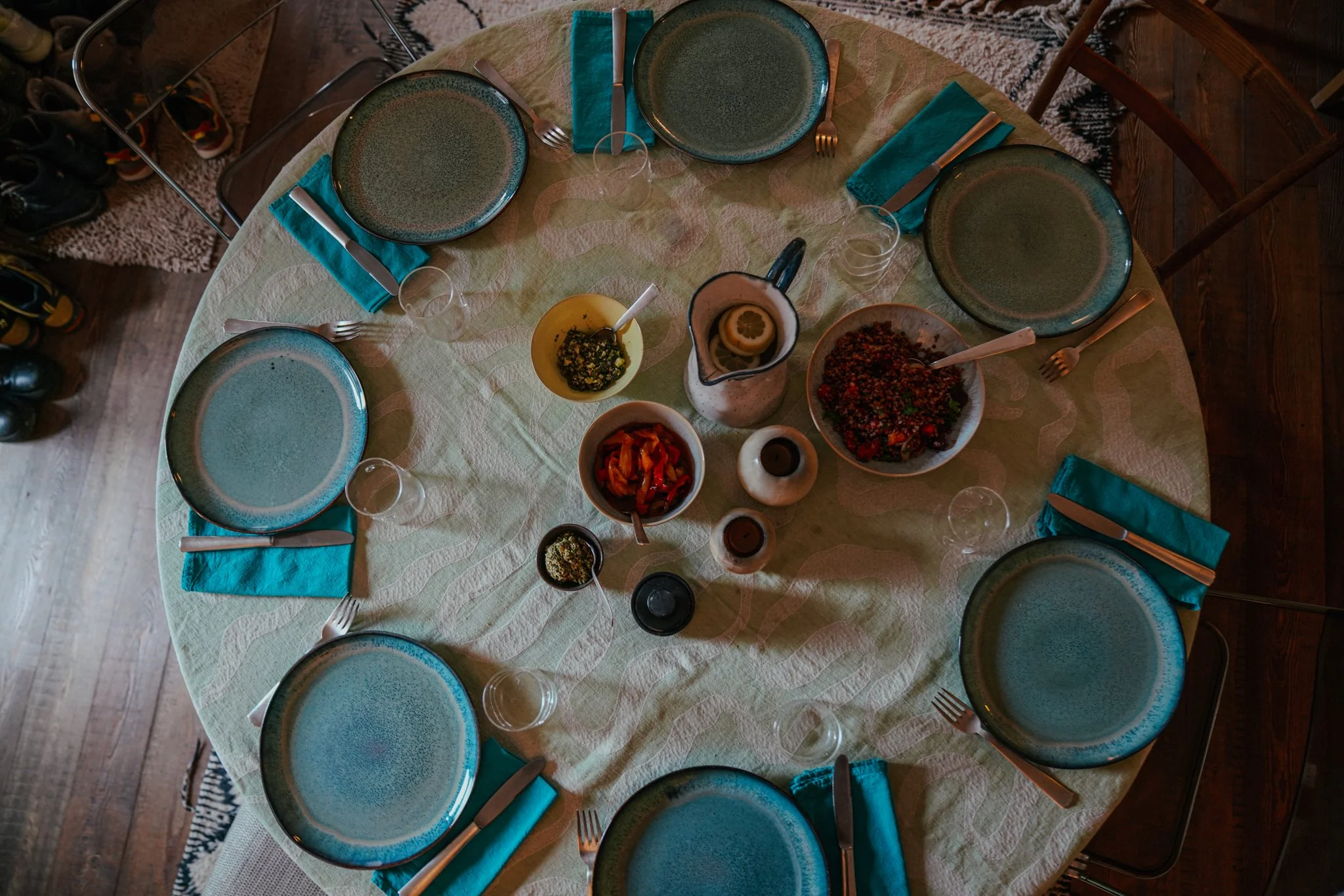 Overhead view of a round dining table set for six with blue plates and teal napkins. The center of the table features assorted dishes, including bowls of various foods and a jug with lemon slices.