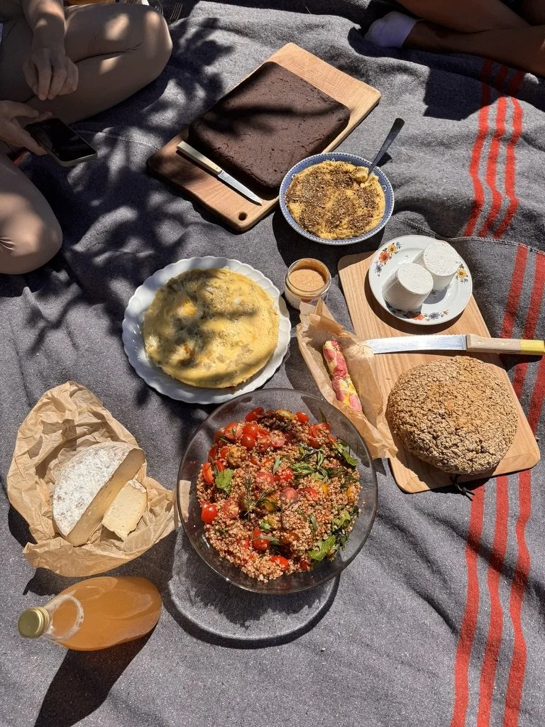 Picnic spread with salad, cheese, bread, cake, dessert, and drinks on a blanket under shadows of leaves.