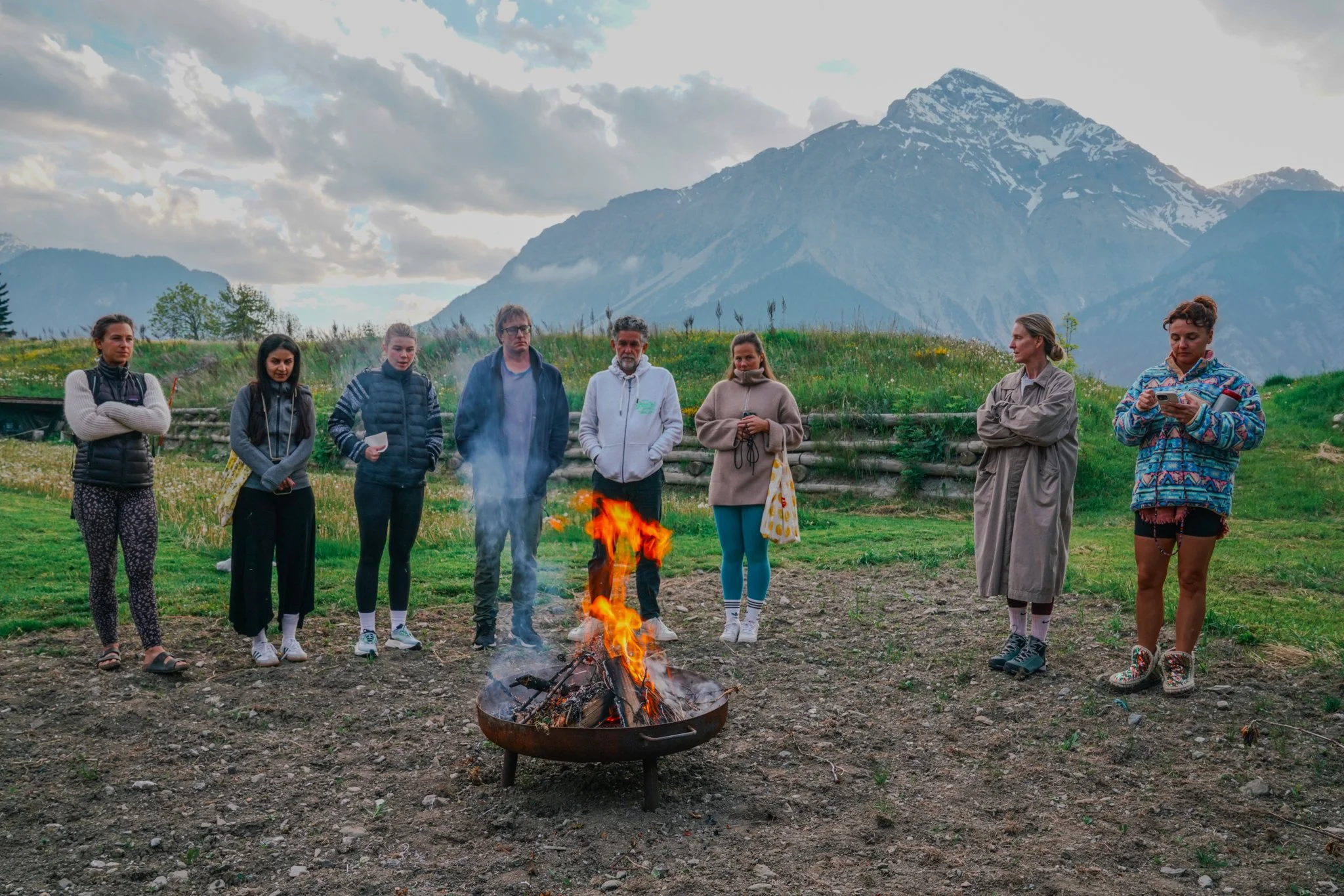 Group of people standing around a campfire in a mountainous landscape.