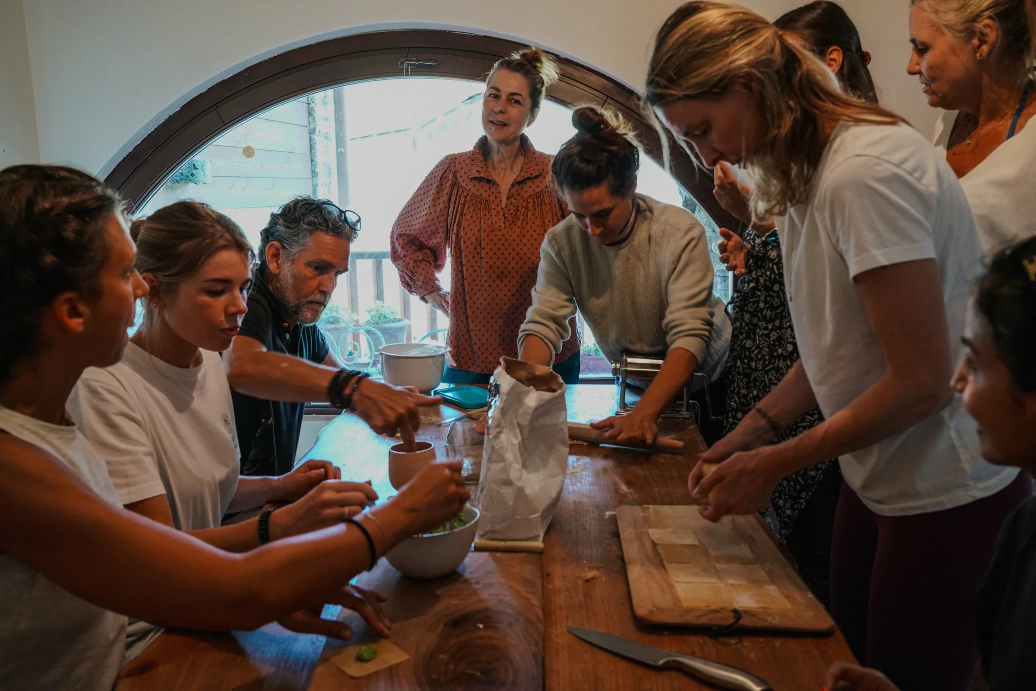 Group of people cooking together at a wooden table, using dough and various ingredients.