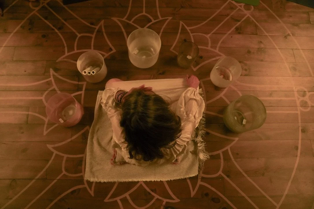 Top view of a person kneeling on a mat surrounded by crystal singing bowls on a wooden floor with an intricate design.