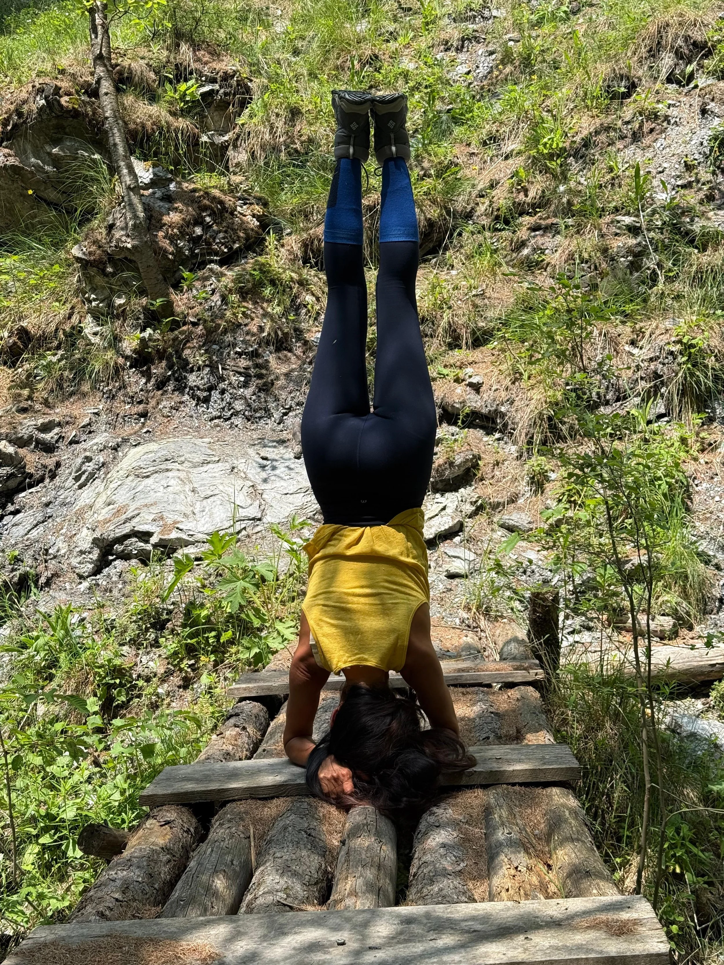 A person doing a headstand on a wooden bridge in a forested area with green plants and rocks.