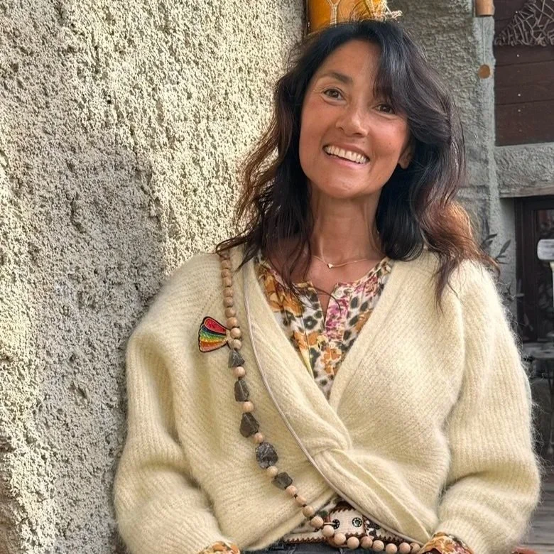 A woman with wavy dark hair is smiling while leaning against a textured wall. She wears a cream sweater over a floral patterned shirt, a necklace, and accessories including a name tag and a beaded bag strap.
