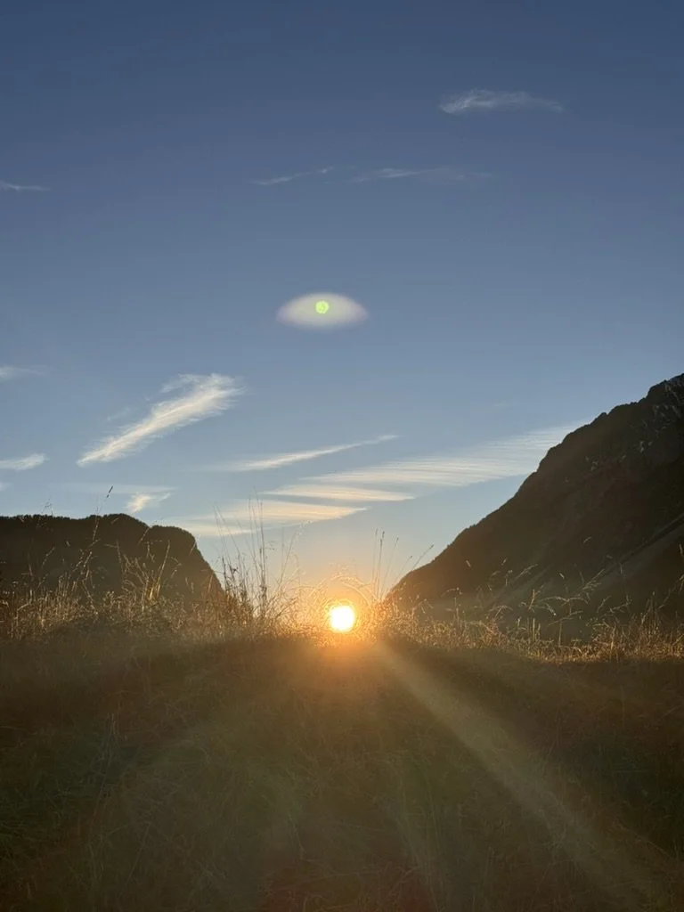 Sunset in a mountainous landscape with grassy foreground, blue sky, some clouds, and a bright sun near the horizon.