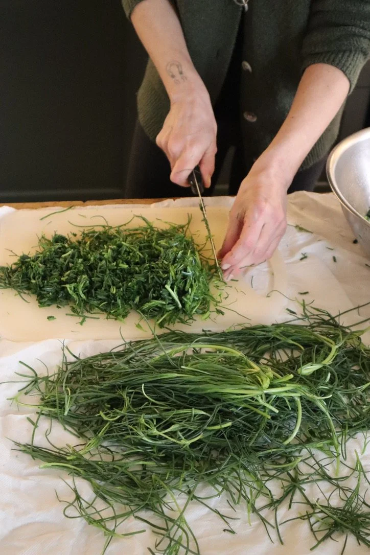 Person chopping fresh green herbs on a wooden cutting board covered with parchment paper, with additional herbs laid out on a white cloth.