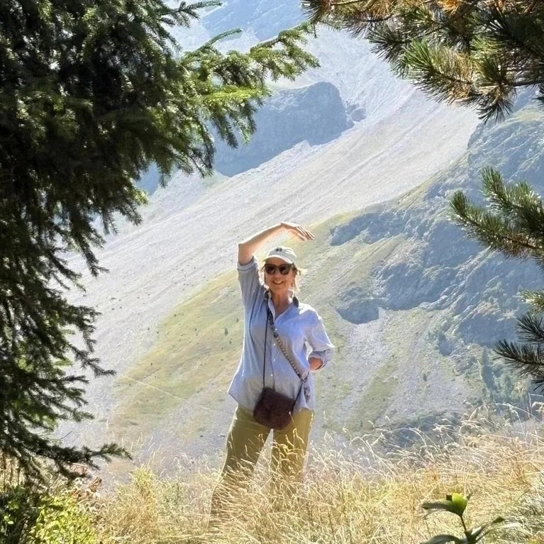 A woman standing outdoors in a mountainous area, smiling, wearing a hat and sunglasses, with trees and mountain slopes in the background.