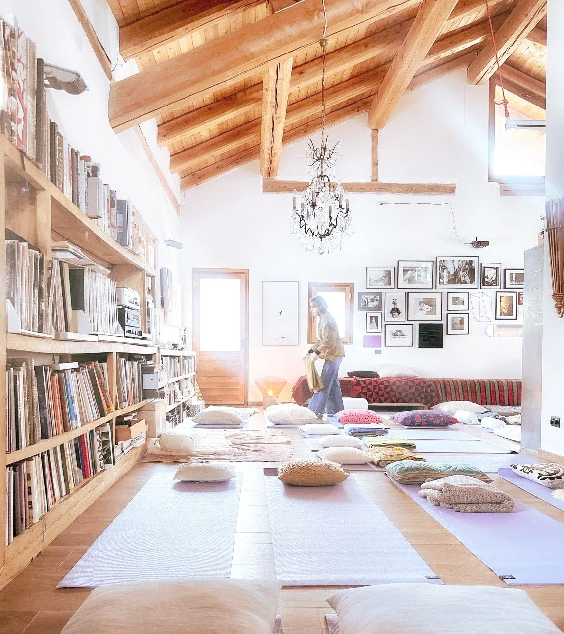 A cozy, well-lit living room with a wooden vaulted ceiling, bookshelves filled with books on the left, a white wall decorated with framed photos, and a woman standing among cushions and mats on the floor preparing for a yoga or meditation session.