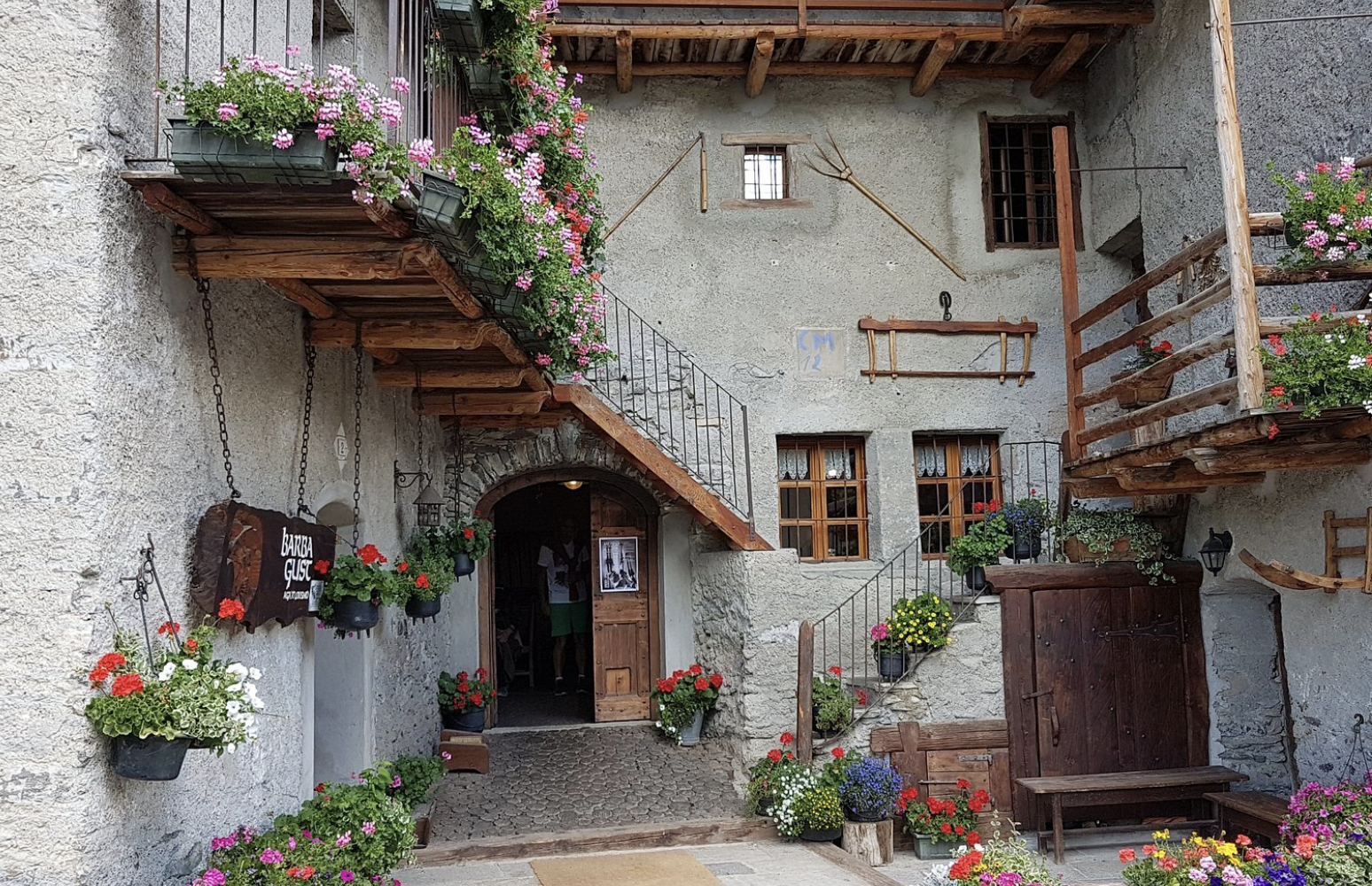 A rustic European courtyard with stone walls, wooden balconies, and colorful potted flowers. There is an arched wooden doorway and small windows with curtains, along with hanging flower pots and wooden outdoor furniture.