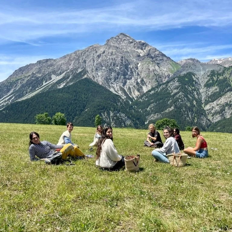 A group of people sitting on a grassy field with mountains in the background, enjoying a picnic.
