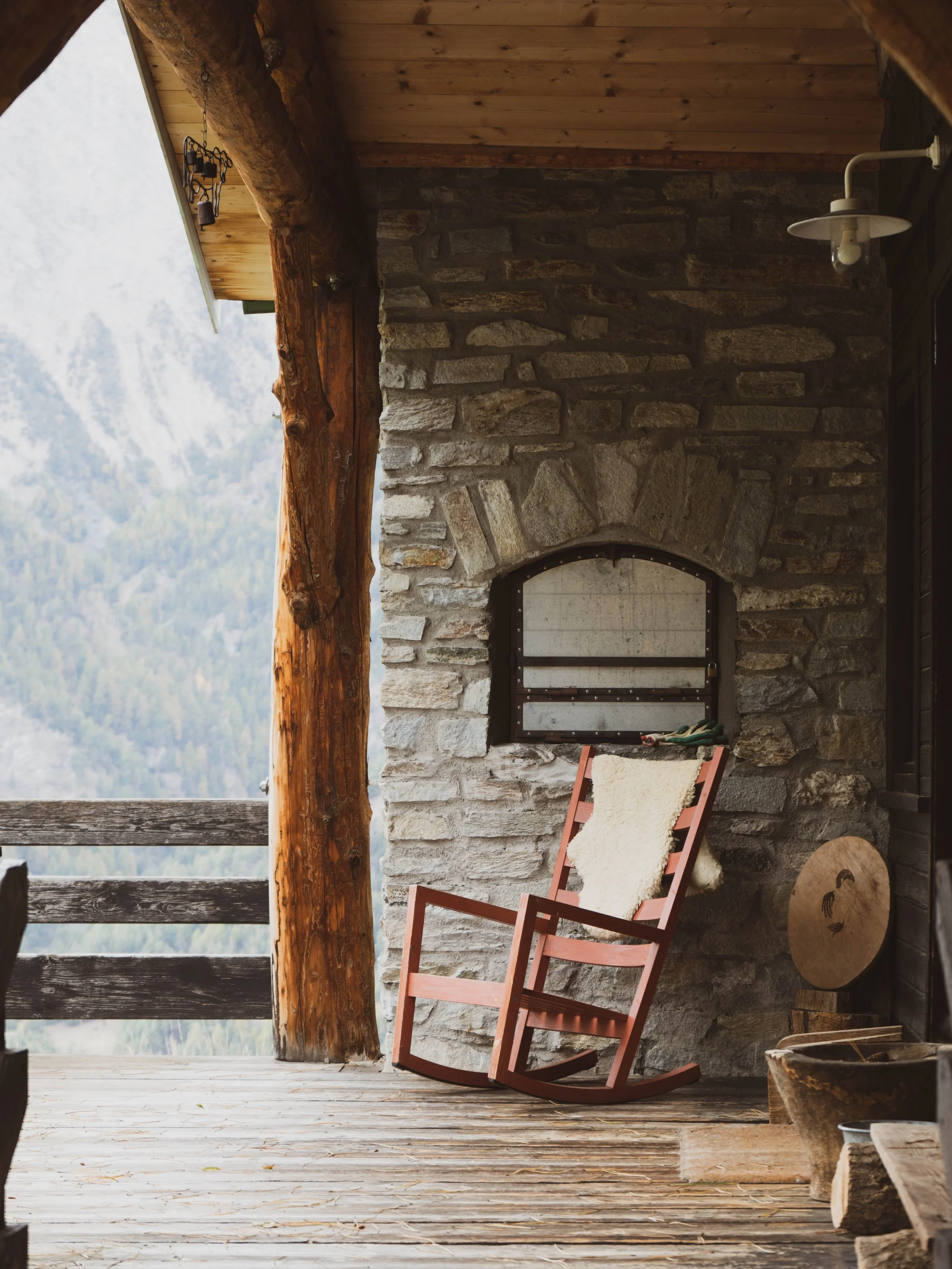 A porch with a wooden rocking chair with a white sheepskin cushion, against a stone wall with a window, overlooking a mountain landscape.