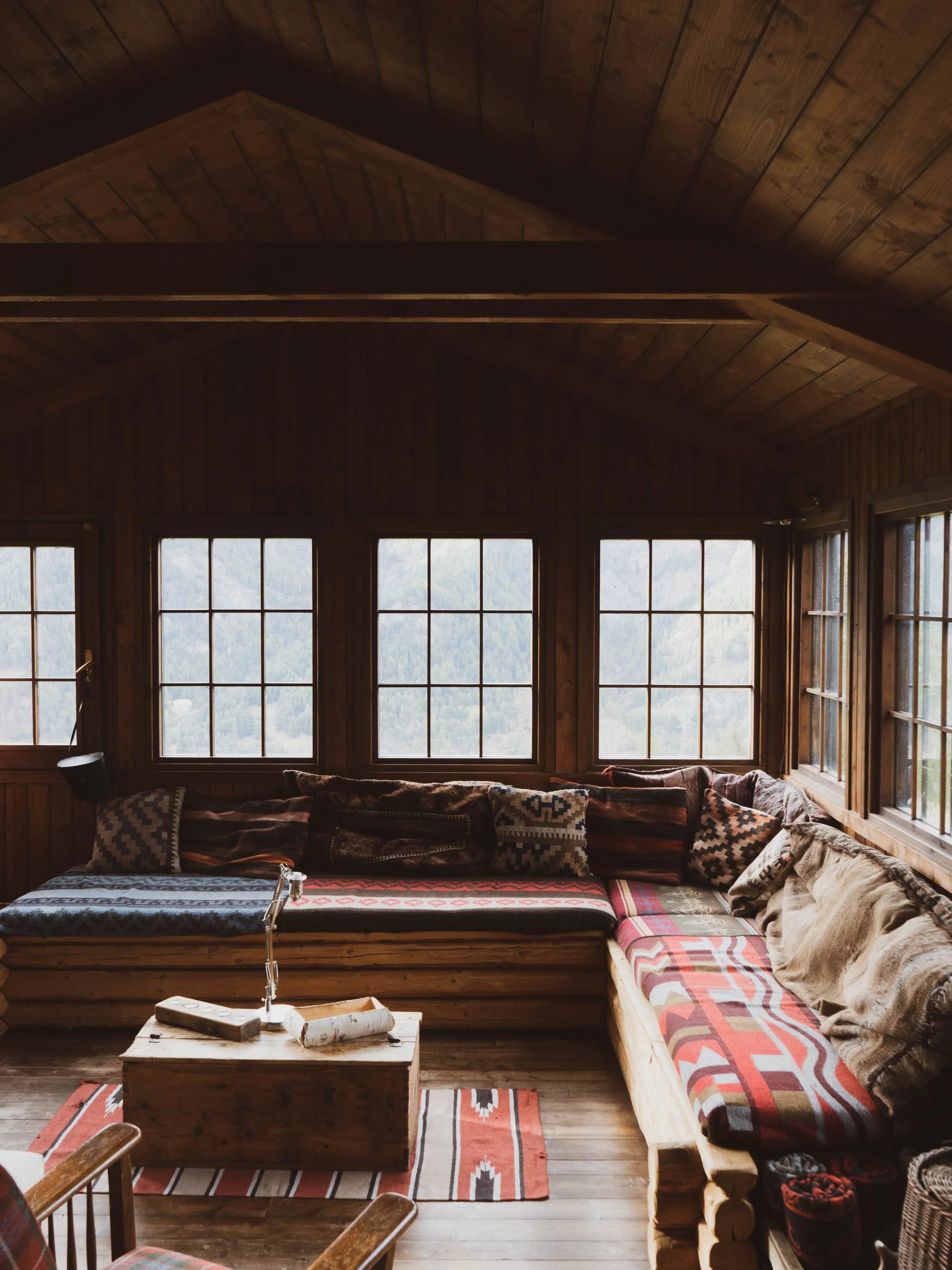 A cozy living room with wood-paneled walls and ceiling, featuring a corner sectional sofa with patterned cushions, a wooden coffee table with logs, and large windows showing a mountain landscape outside.