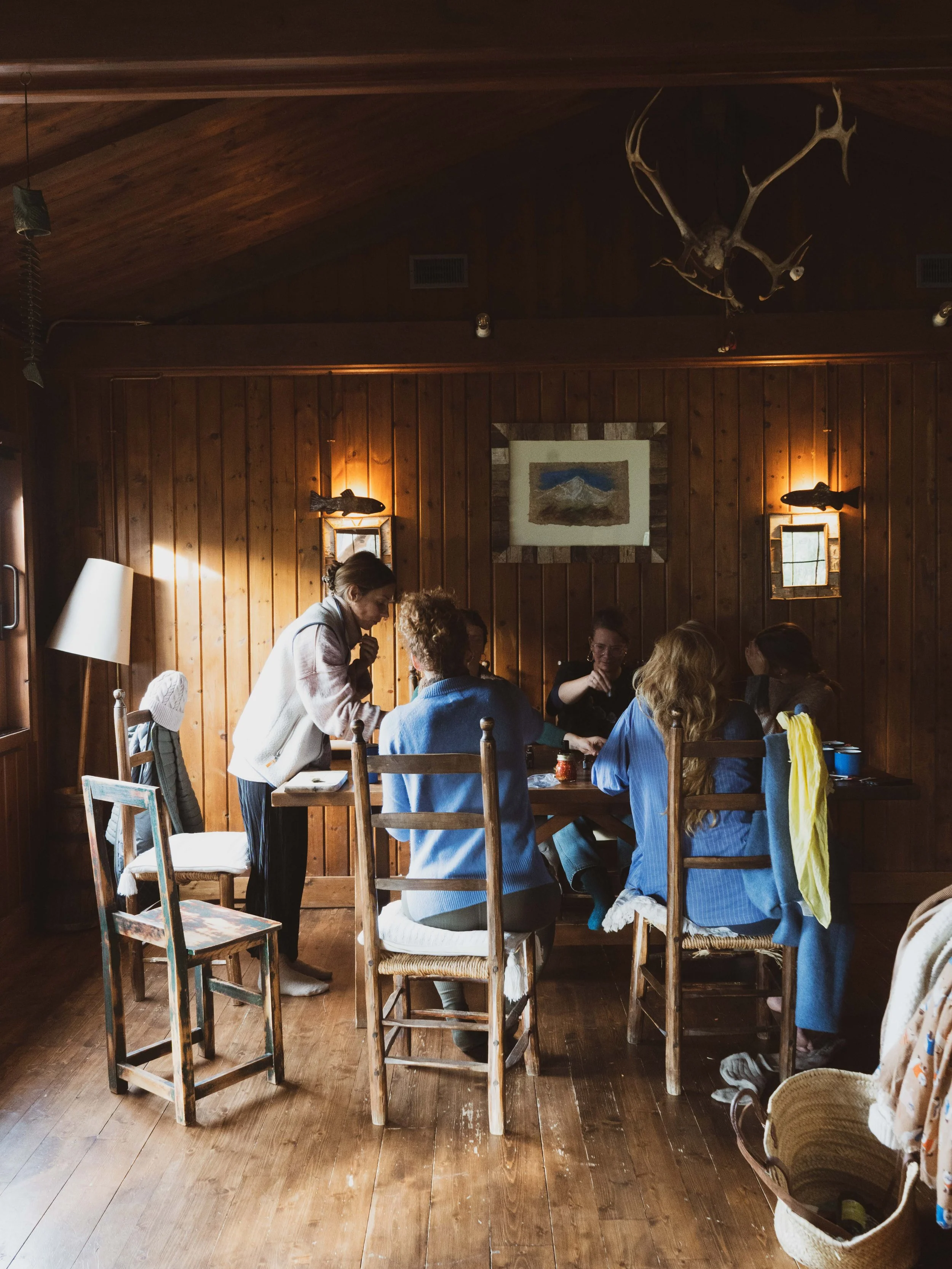 A group of women gathered around a wooden table in a rustic room with wood-paneled walls, some wearing blue jackets. A woman stands and leans over to speak to one of the seated women. The room has deer antlers mounted on the ceiling, warm lighting, and a framed picture of a mountain landscape on the wall.