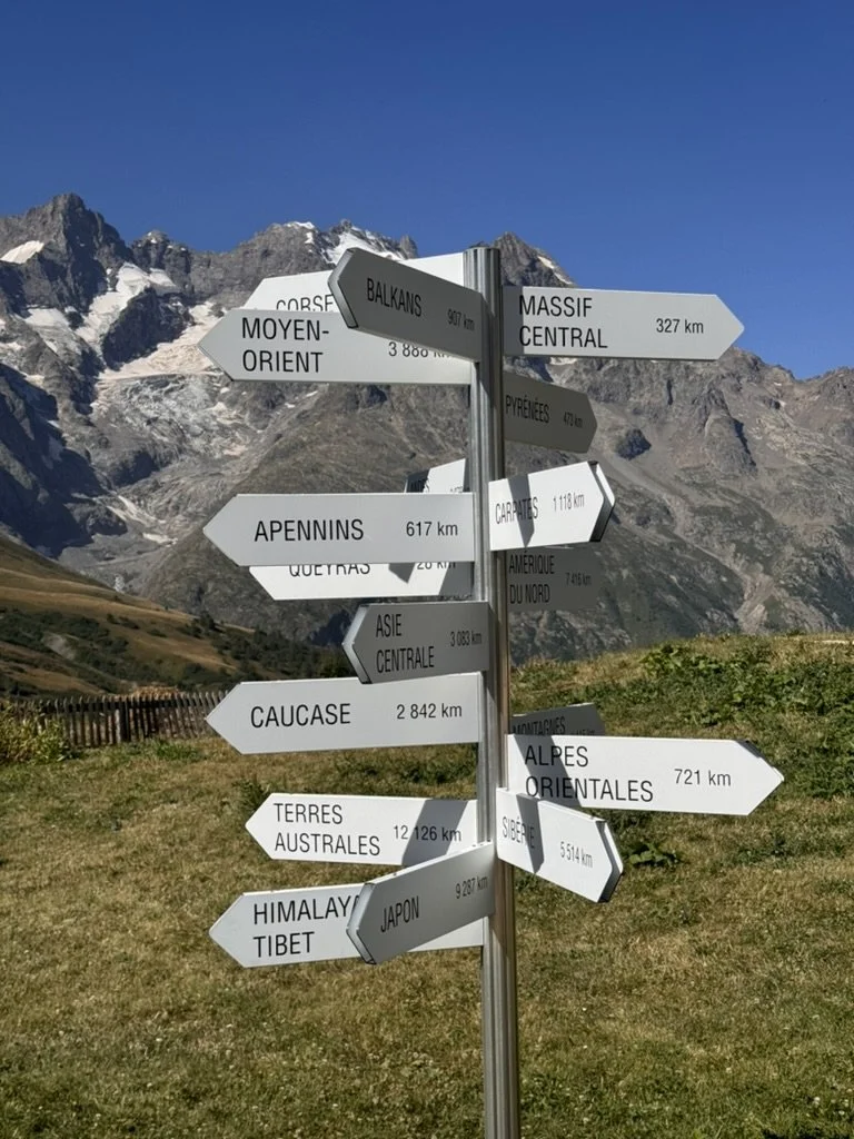 A signpost with multiple directional signs in French, pointing to various mountain ranges and locations, set against a mountain landscape and blue sky.