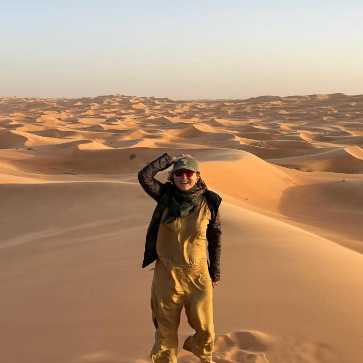 A smiling woman in a desert with sand dunes, wearing sunglasses, a beige jumpsuit, a black jacket, and a green cap, saluting with her right hand.