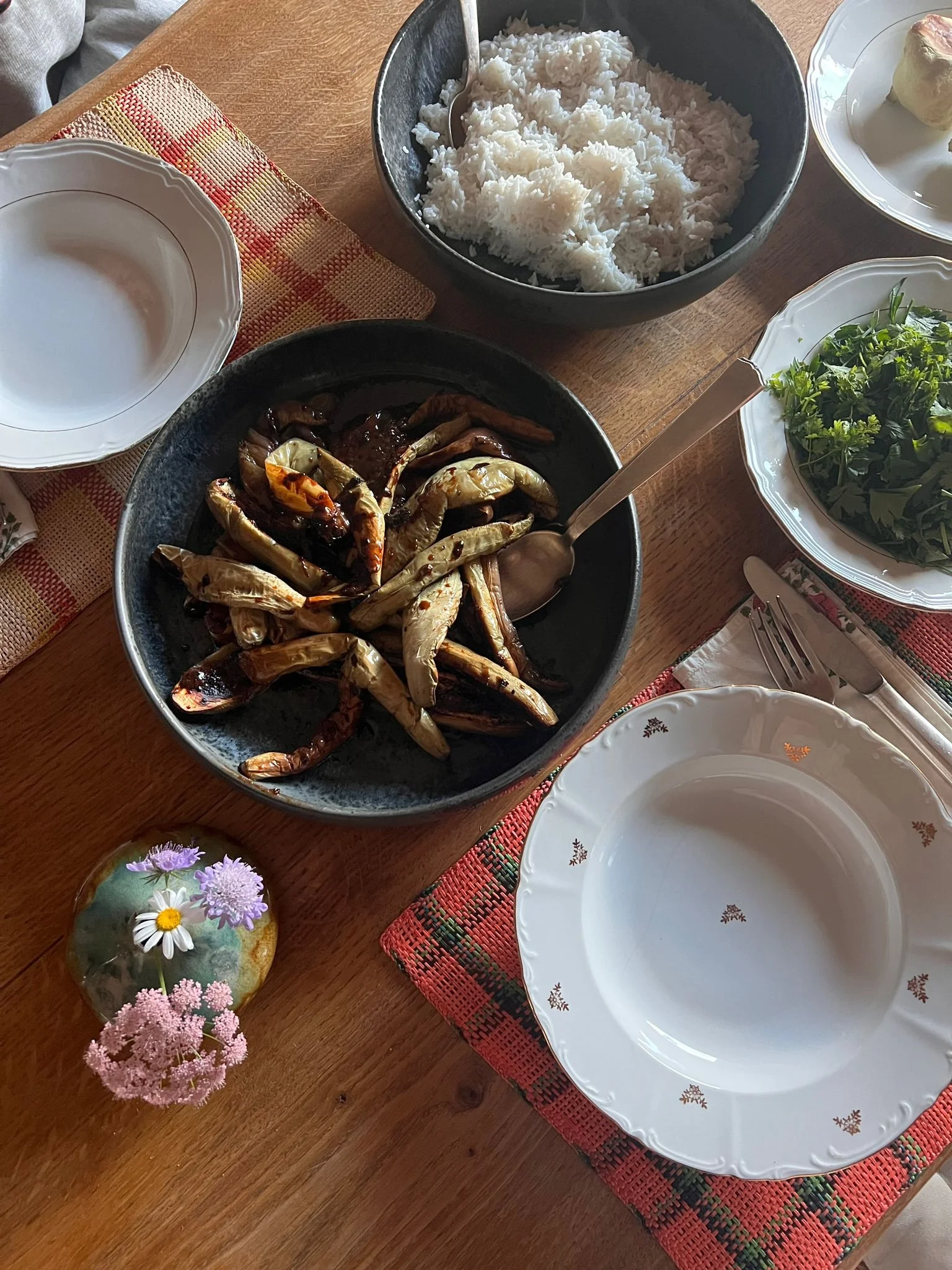 A wooden dining table set with a white empty plate, a bowl of cooked rice, a bowl of grilled eggplant, a small vase of pink and white flowers, and a bowl of fresh greens.