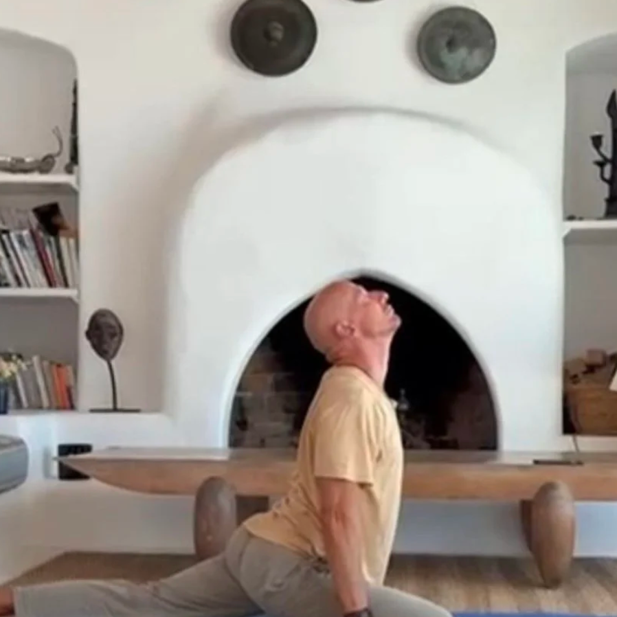A man is practicing yoga in a split pose in front of a fireplace in a living room, looking upwards.
