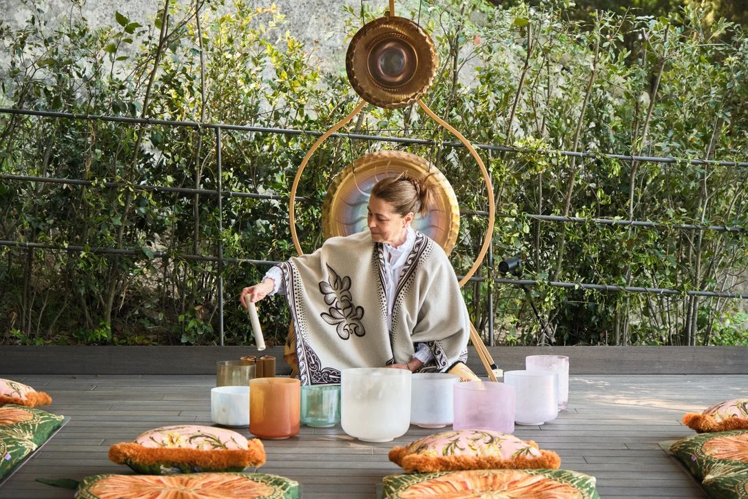Larissa Giers playing crystal singing bowls outdoors at the Belmond Hotel Villa San Michel surrounded by pillows, with greenery and a decorative backdrop.