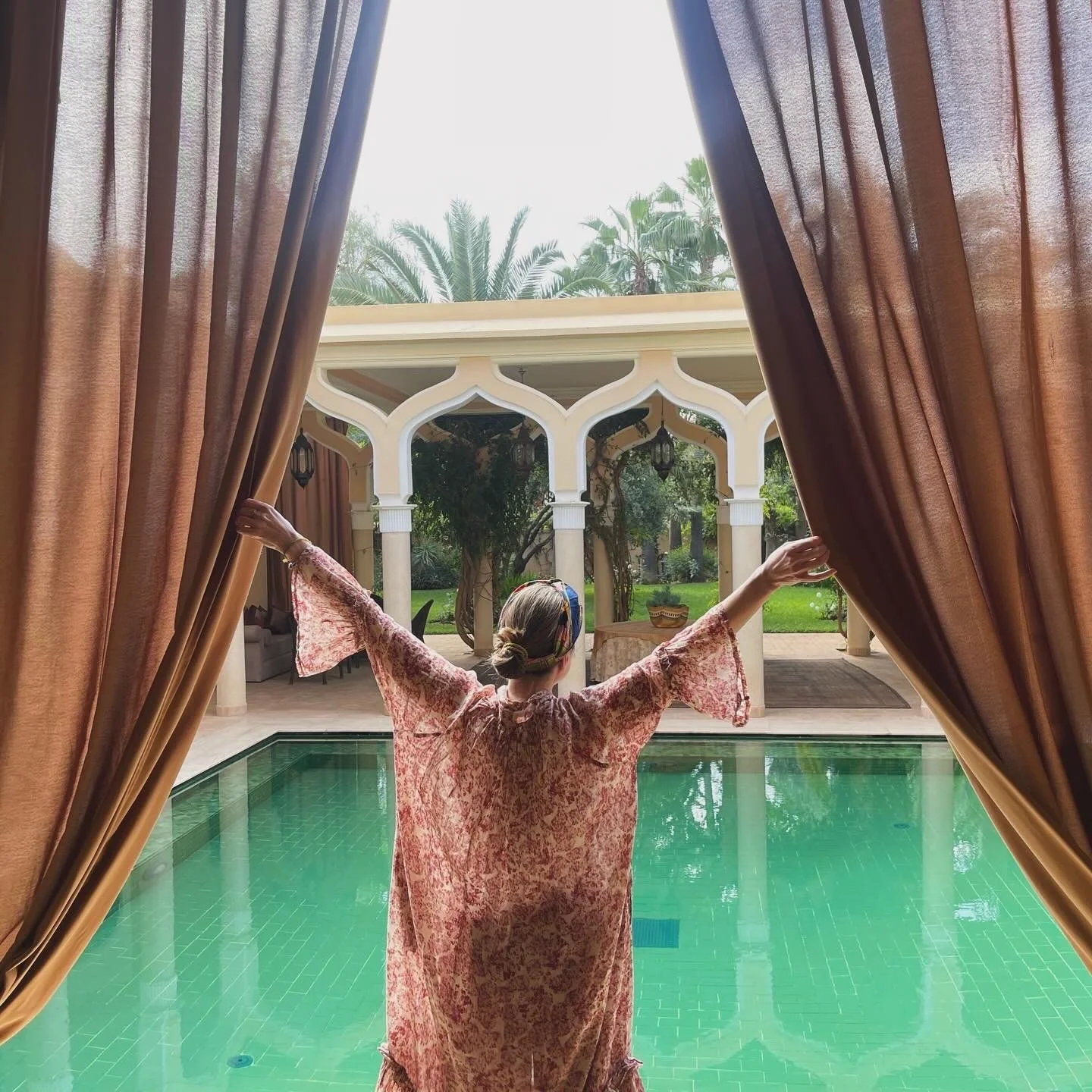 Person standing near pool with arms outstretched, looking out to garden and architectural features beyond, viewed from inside through open curtains.