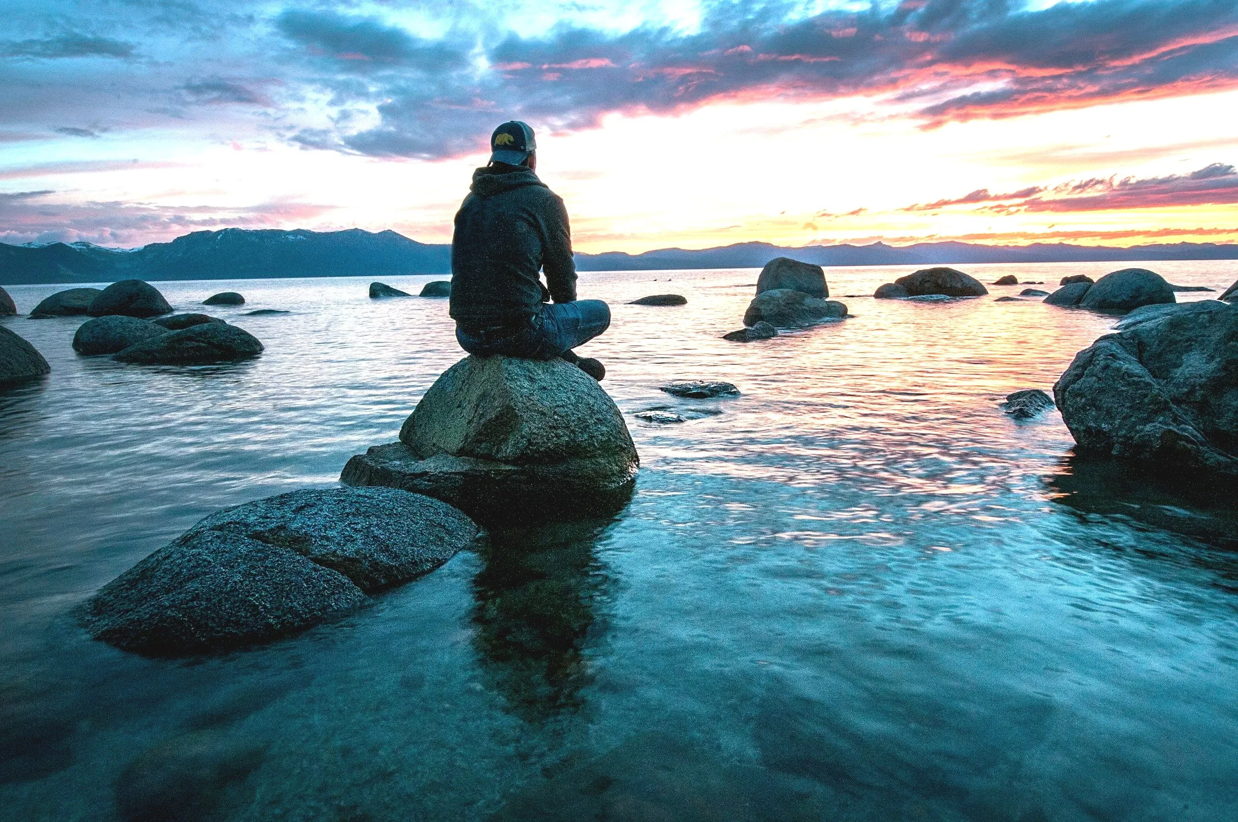 Person sitting on a rock by the water at sunset, with mountains in the background.