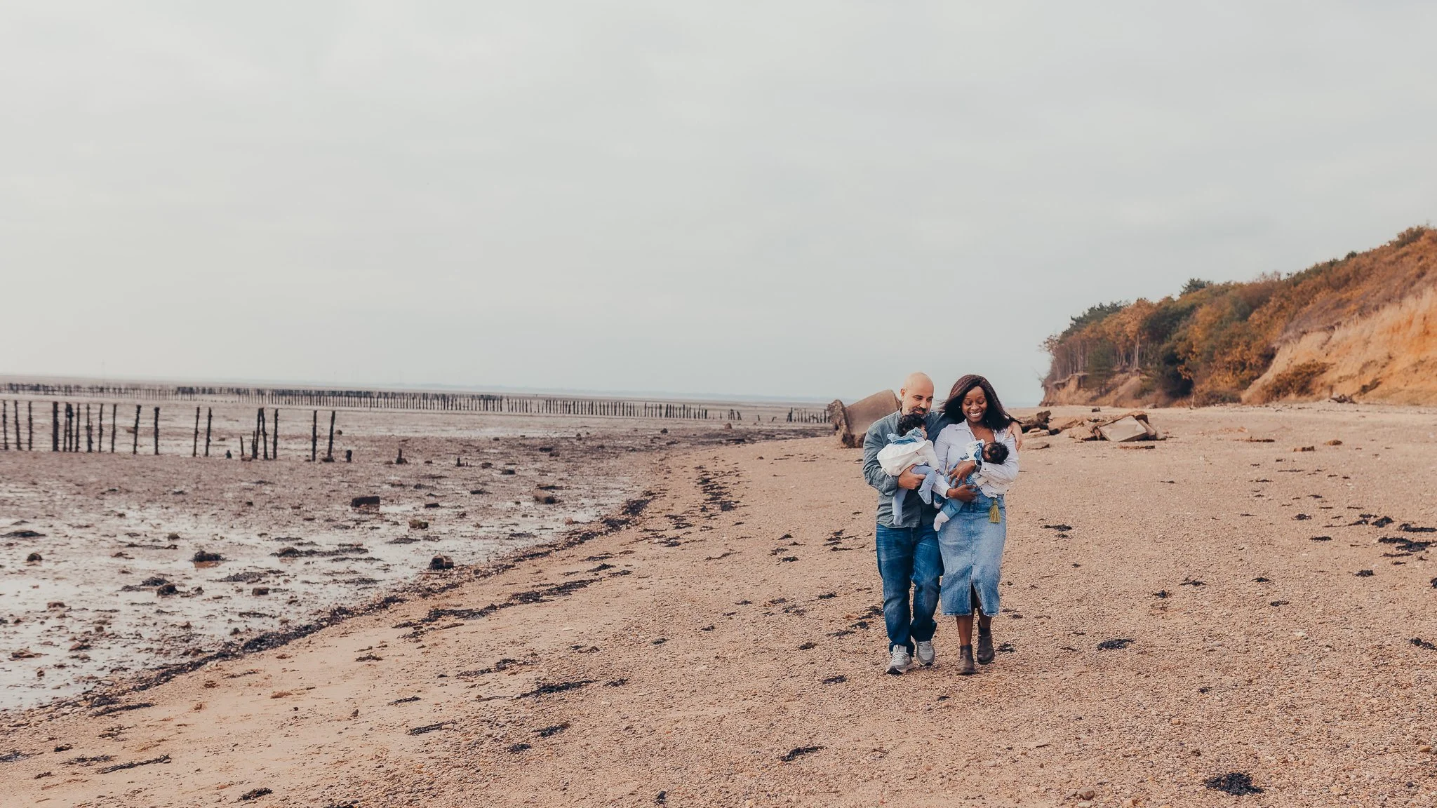 New parents walking with their baby twin girls during a morning family photoshoot at an Essex beach