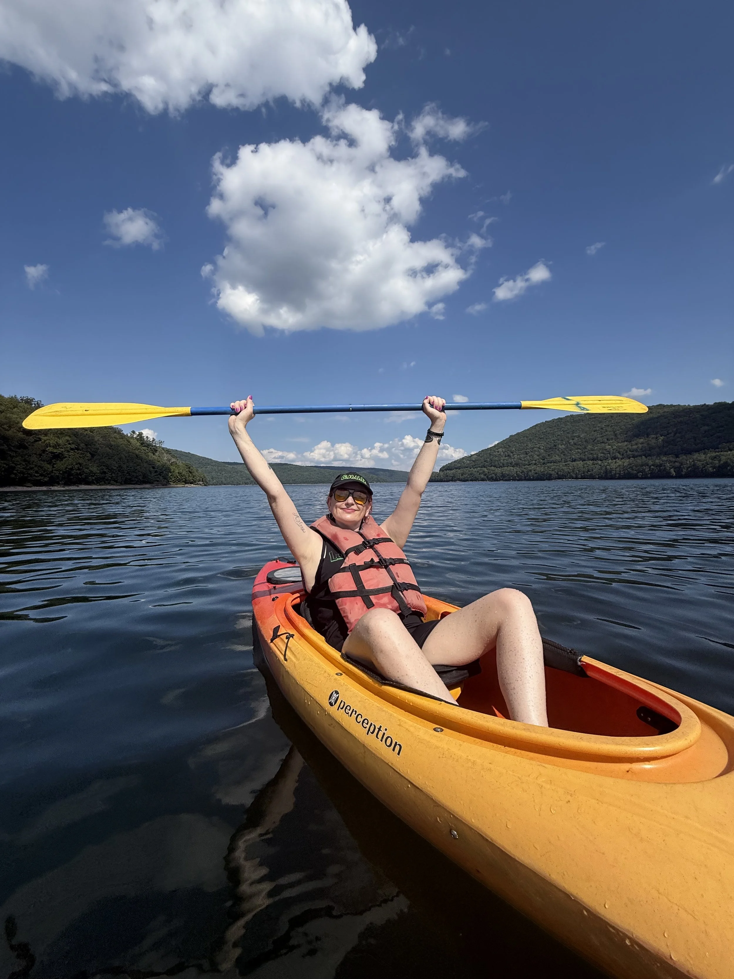 Woman smiling in a kayak on a lake, holding a yellow paddle above her head, with hills and a partly cloudy sky in the background.