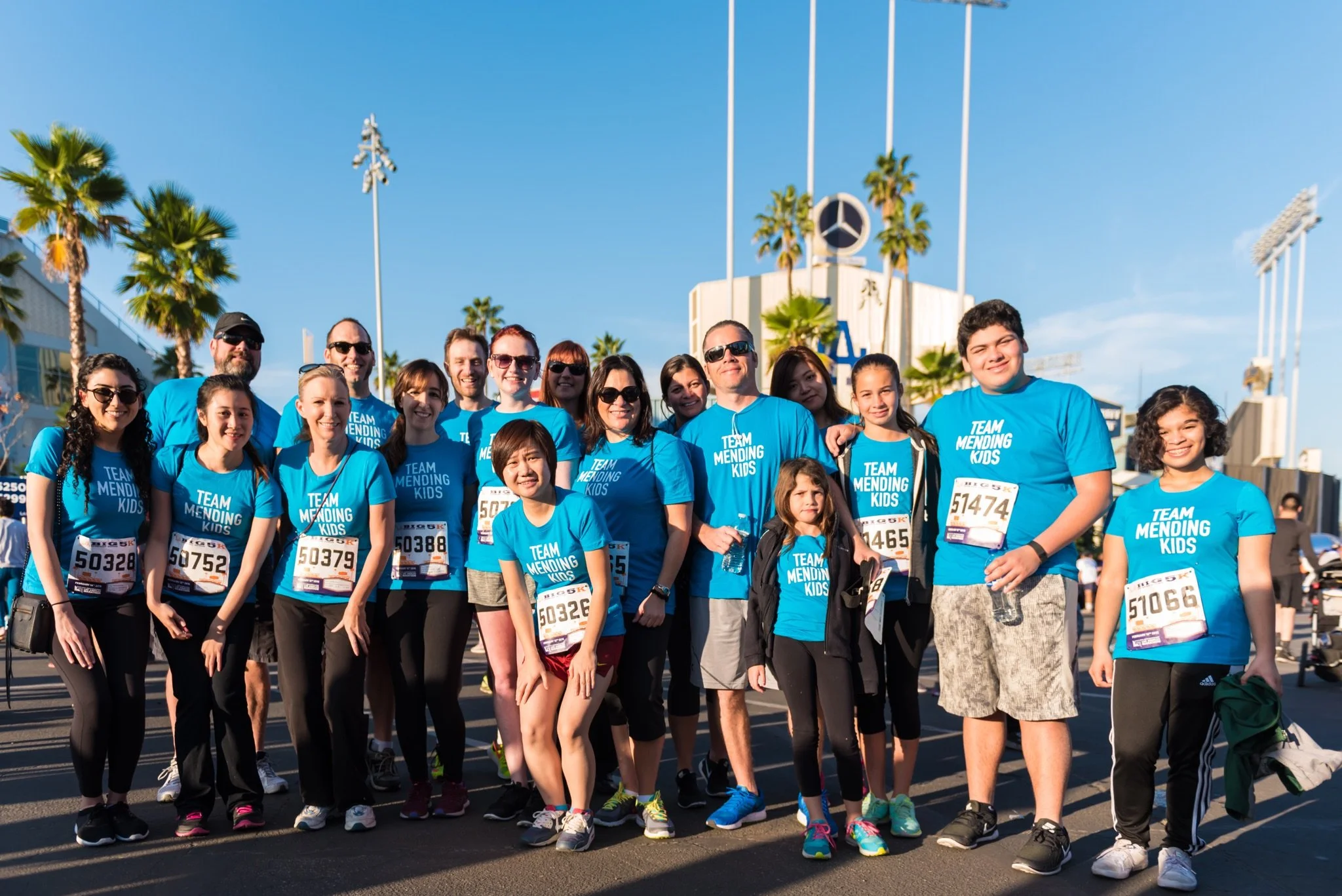A group of people in matching blue shirts with 'TEAM MENDING KIDS' printed on them, participating in a race event, standing outdoors on a sunny day with palm trees and a sports stadium in the background.