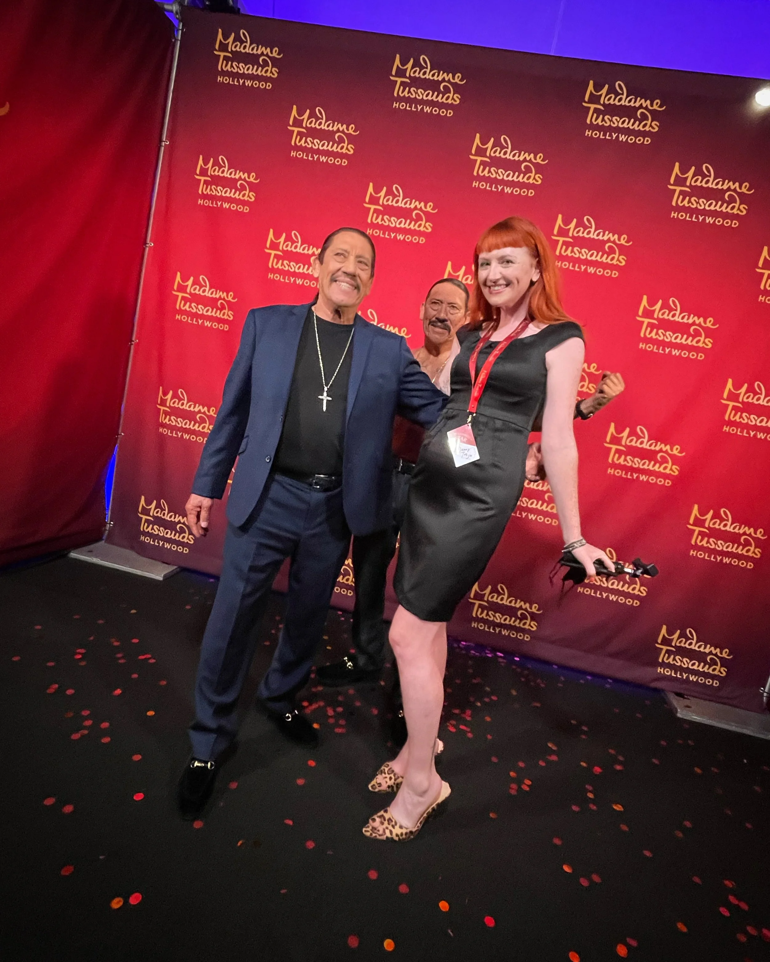Three people posing together at Madame Tussauds Hollywood, with a red backdrop featuring the Madame Tussauds logo repeated multiple times. The group includes a man in a dark blue suit with a cross necklace, a woman with red hair in a black dress, and another man in the background wearing glasses and a dark suit. The woman is smiling, and the men are making gestures. The floor is decorated with red and yellow confetti.