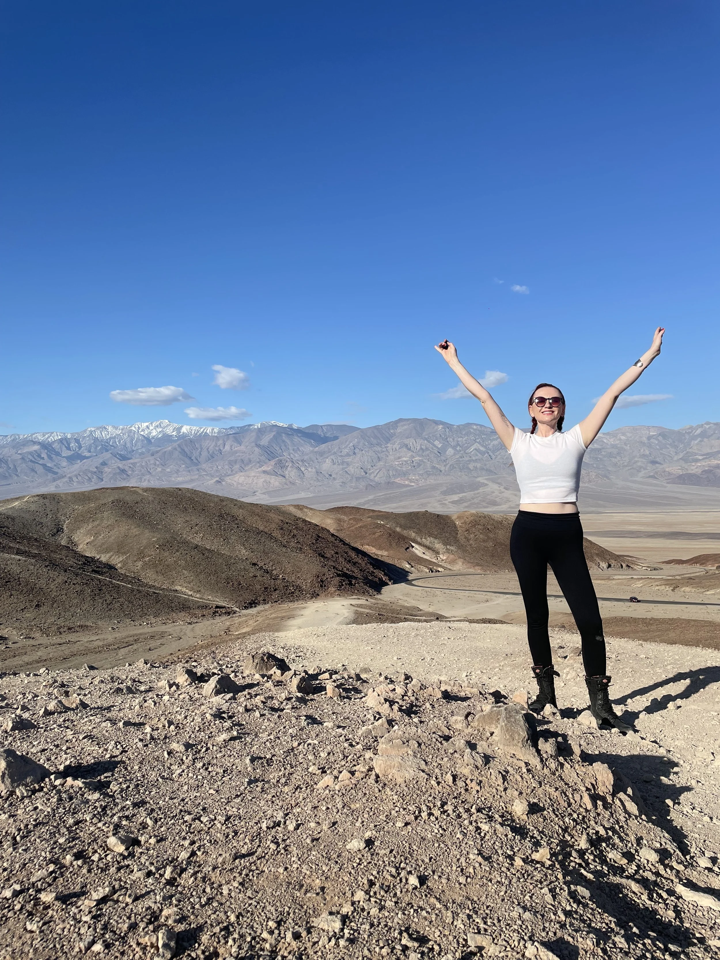 A woman standing on rocky terrain in a desert landscape with mountains in the background, arms raised in celebration, wearing sunglasses, a white crop top, black pants, and hiking boots.