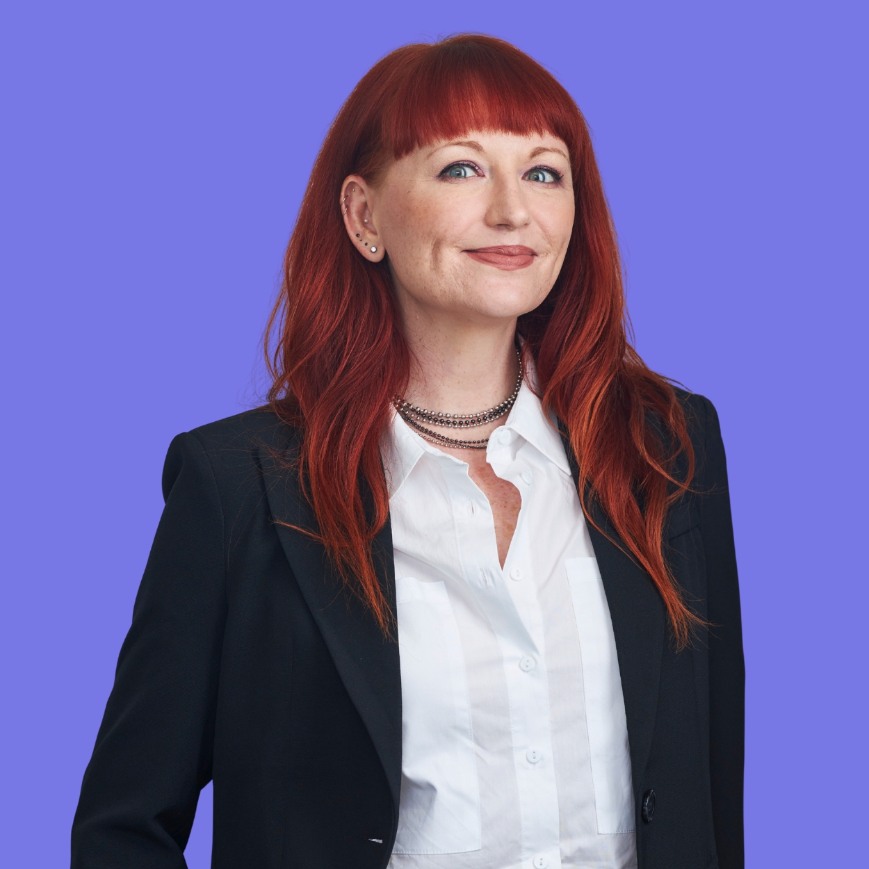 A woman with red hair, wearing a black blazer, white shirt, layered necklaces, and multiple earrings, smiling against a blue background.