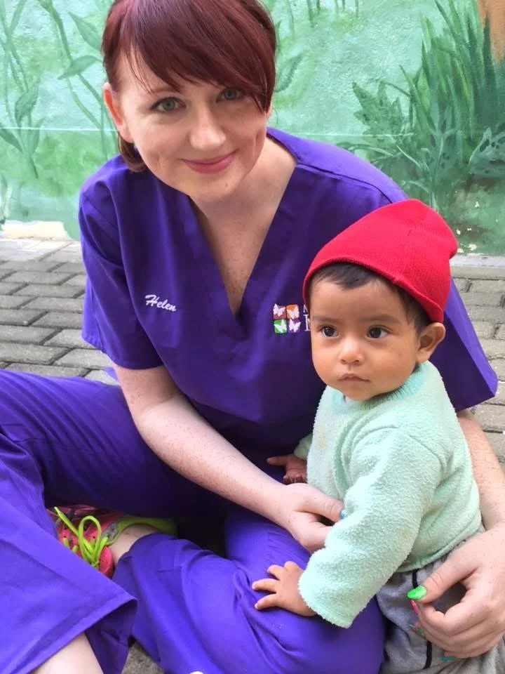 A woman with short red hair wearing purple scrubs, smiling and sitting outside on a paved surface with a young child. The child has short black hair, a red beanie, a light green fleece jacket, and is looking off to the side. The woman is holding the child, and they are in front of a green painted background that appears to be an outdoor mural or wall.
