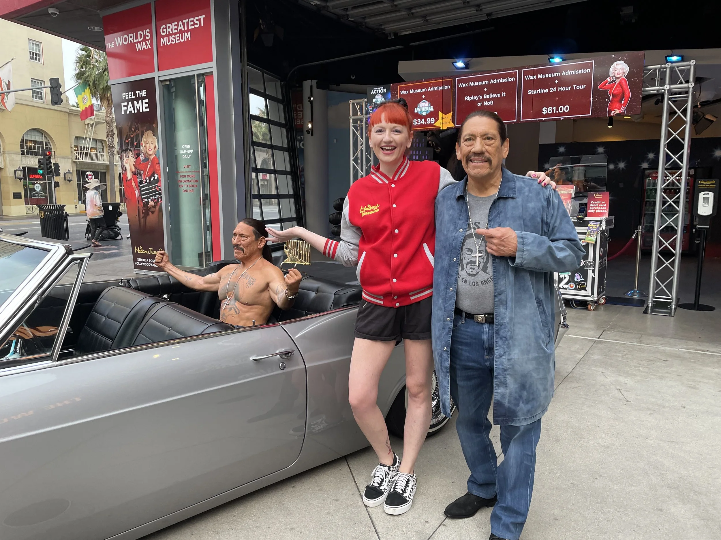 Two people posing outside Madame Tussauds wax museum with life-sized wax figure of a man in a convertible car. The woman is wearing a red and gray jacket, black shorts, and sneakers, smiling and gesturing toward the wax figure. The man is wearing a gray T-shirt, blue jeans, and a denim jacket, smiling and pointing at the wax figure.
