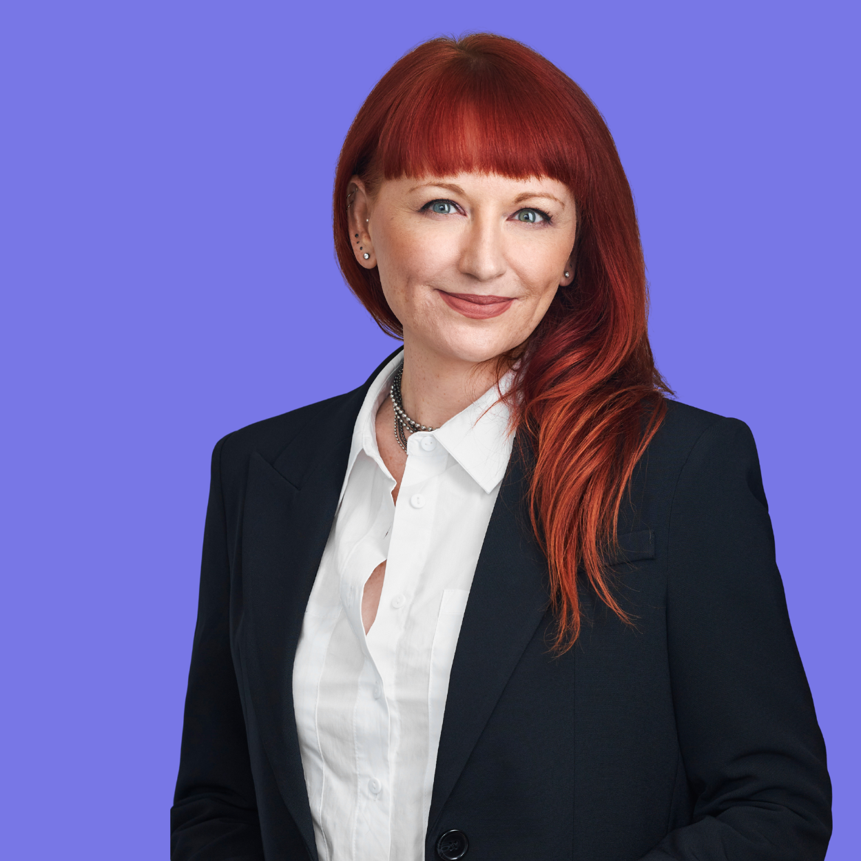A woman with red hair, wearing a black blazer and white shirt, smiling at the camera against a purple background.