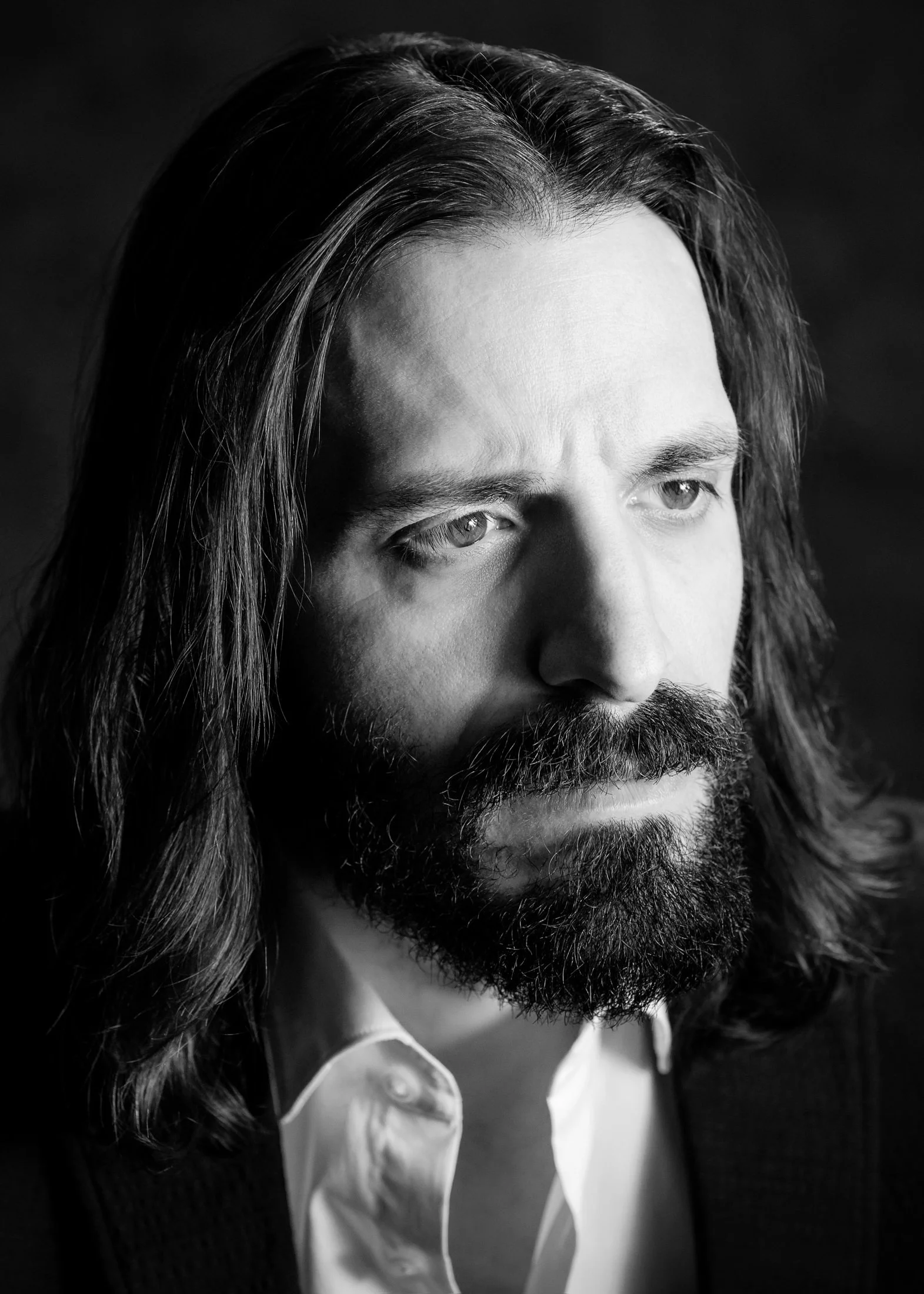 Black and white portrait of a man with long hair and a full beard, wearing a shirt and blazer, looking contemplative.