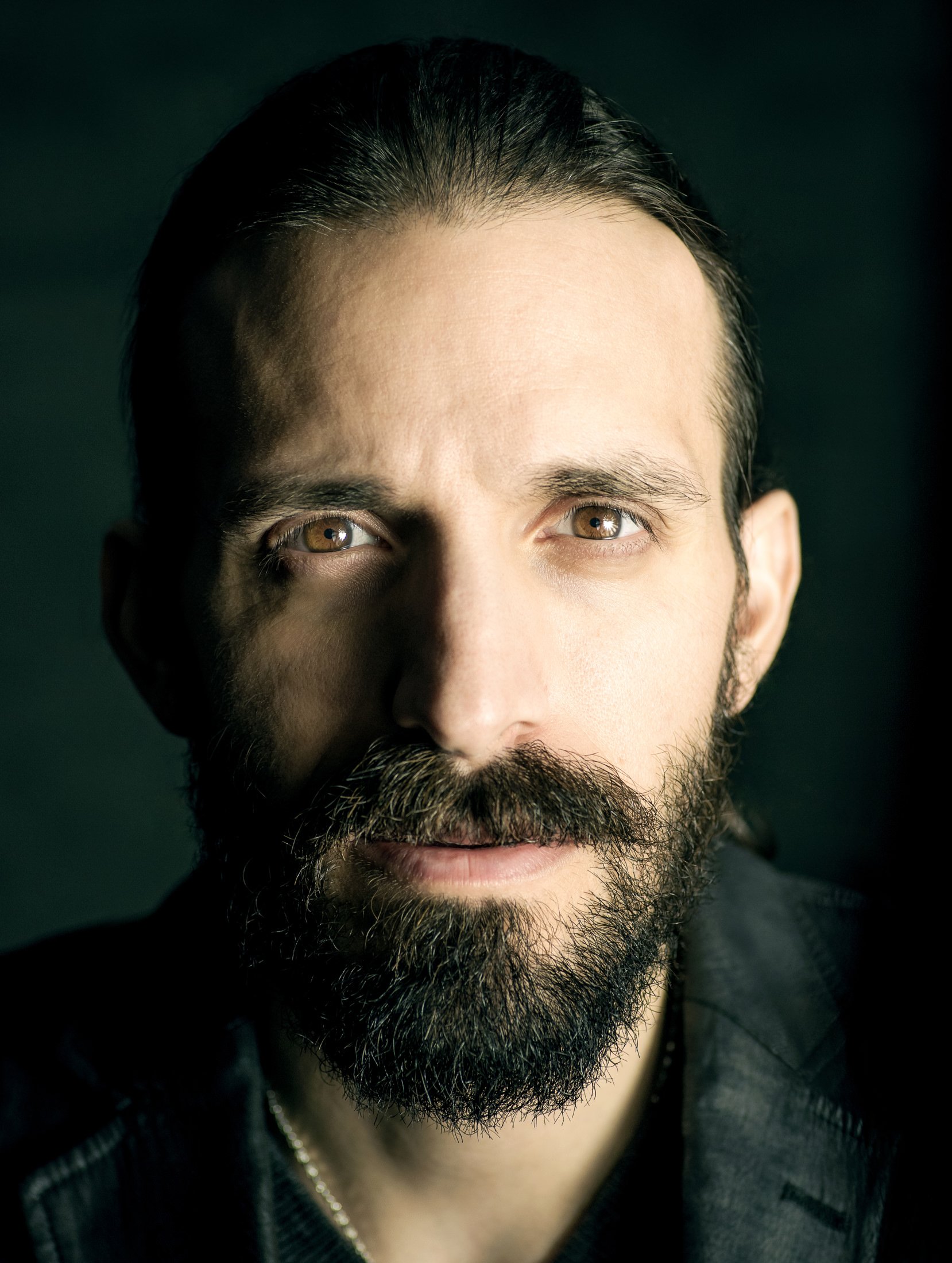 Close-up portrait of a man with dark hair, brown eyes, a full beard, and mustache, wearing a dark jacket, against a dark background.