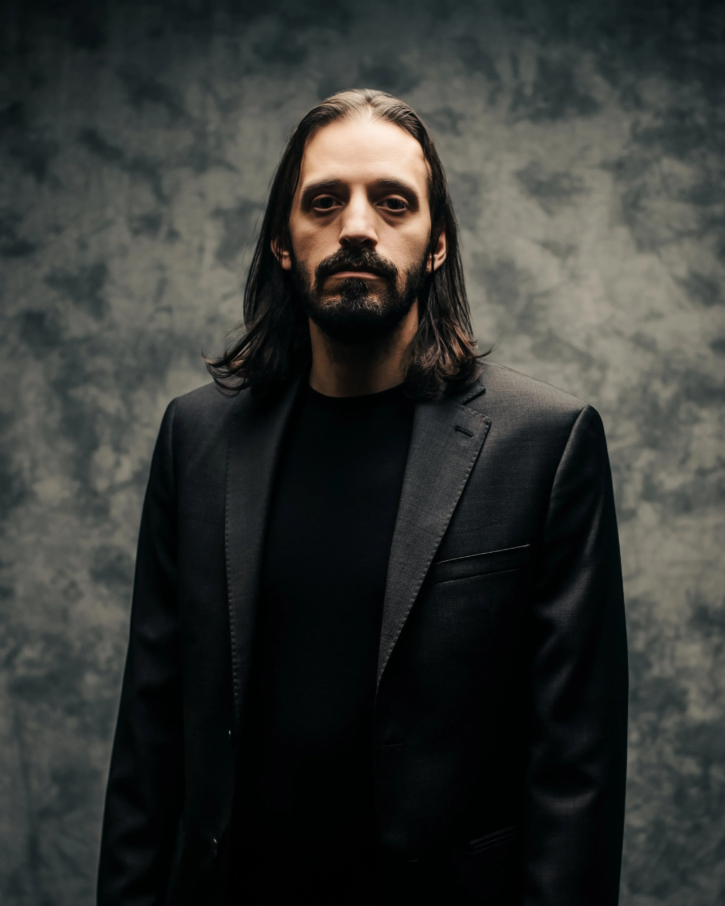 A man with long dark hair and a beard, wearing a black blazer and black shirt, standing against a textured gray background.