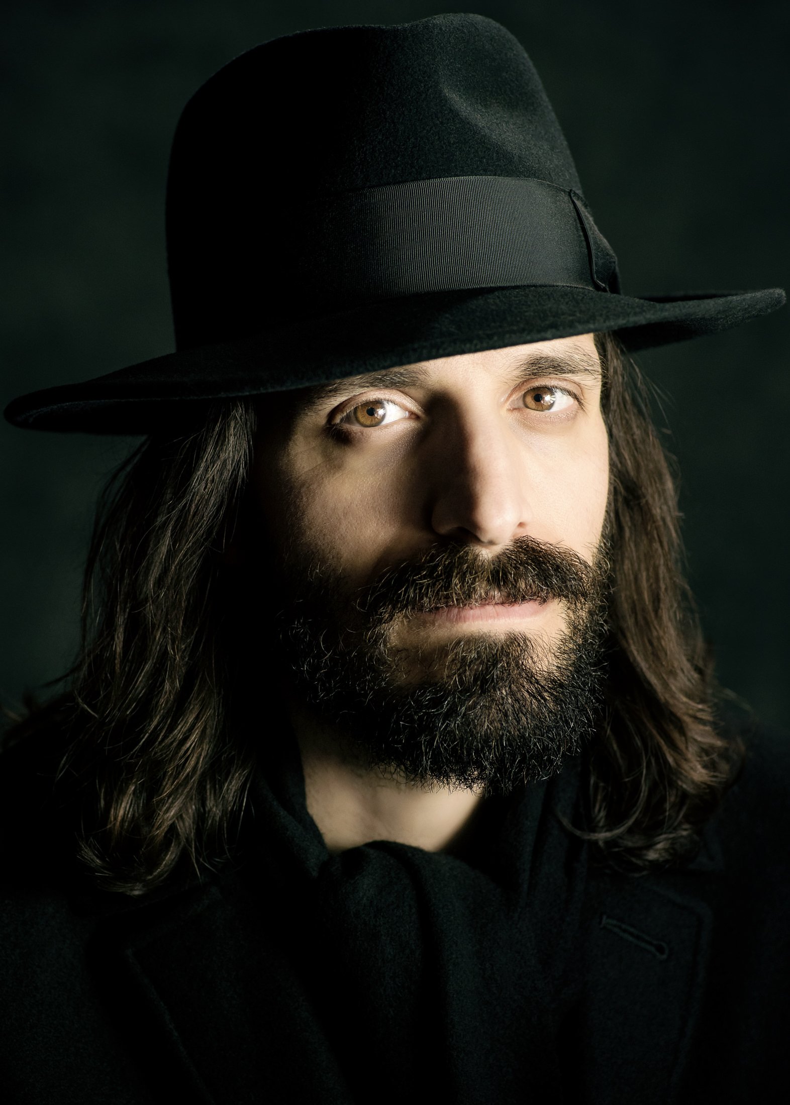 A man with long hair and a beard wearing a black hat with a ribbon, looking directly at the camera.