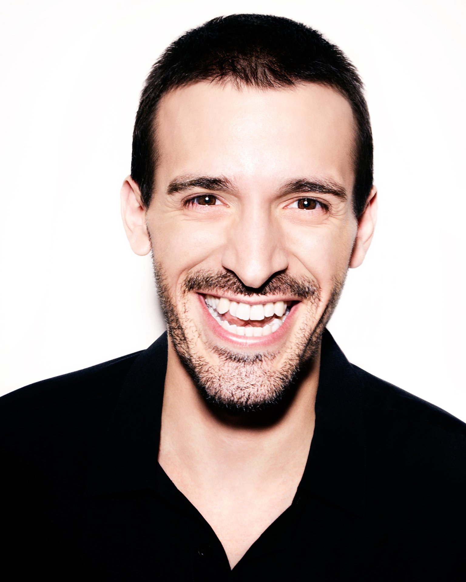 Close-up of a smiling man with short dark hair and a beard, wearing a black shirt, against a white background.
