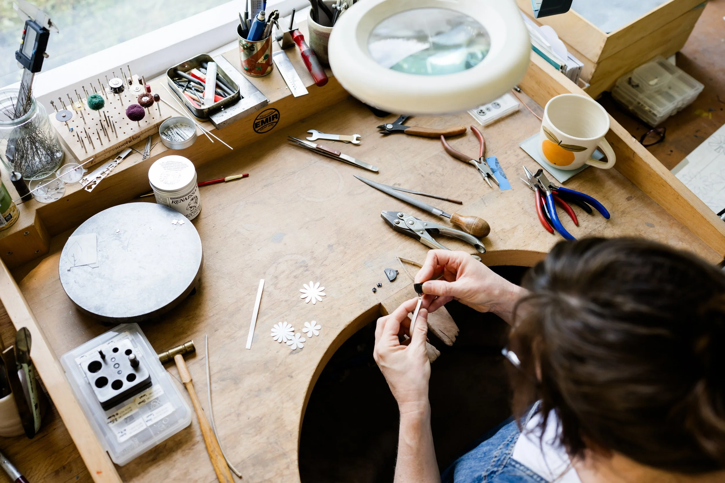 Lindsey working at a jewellers workbench with various tools, small parts, and a magnifying lamp.