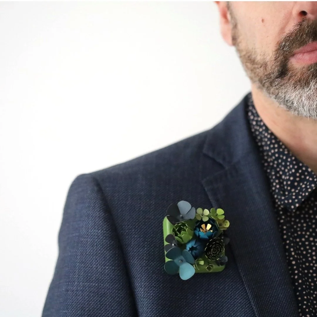 Close-up of a man wearing a dark blue suit with a black and white polka dot shirt. He has a gray beard and is wearing a decorative lapel pin featuring a cluster of small, colorful flowers.