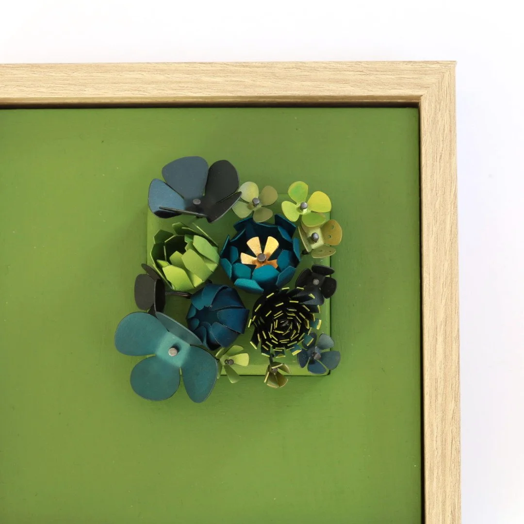 Decorative wall piece with aluminium flowers in shades of blue, green, and black on a green background with a light wooden frame.