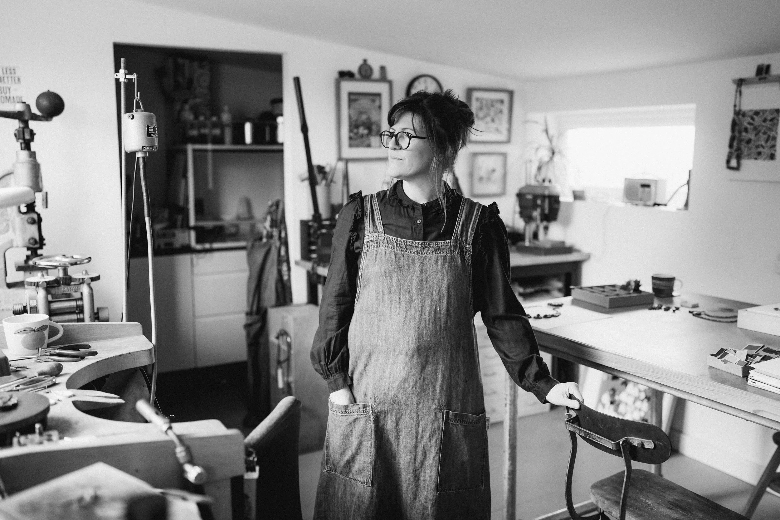 Lindsey, standing in her studio. wearing glasses and a denim apron, surrounded with tools, framed pictures, and art supplies.