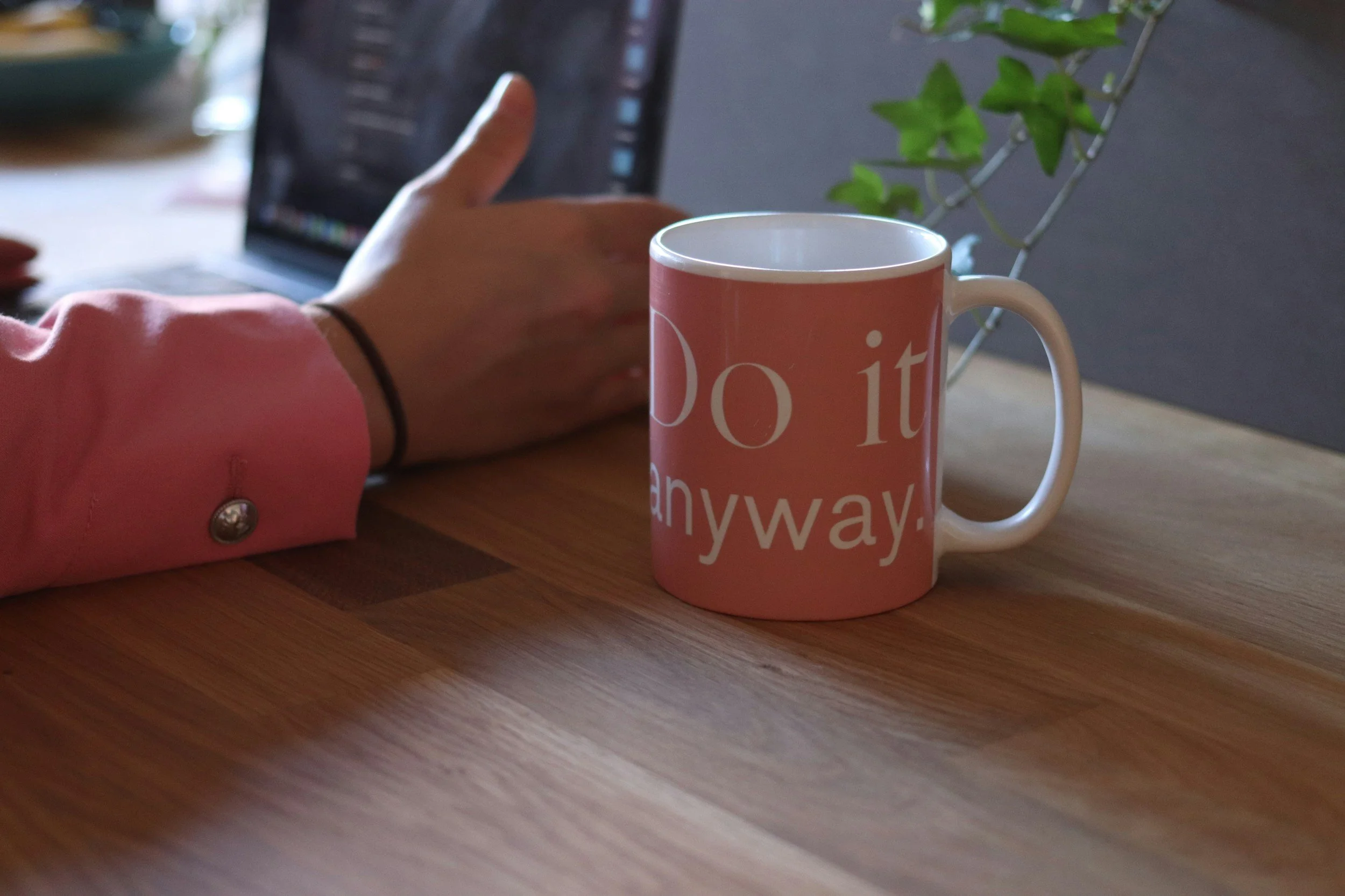 A pink coffee mug with white text that says 'Do it anyway' on a wooden table, with a person's arm and hand reaching towards a laptop in the background.