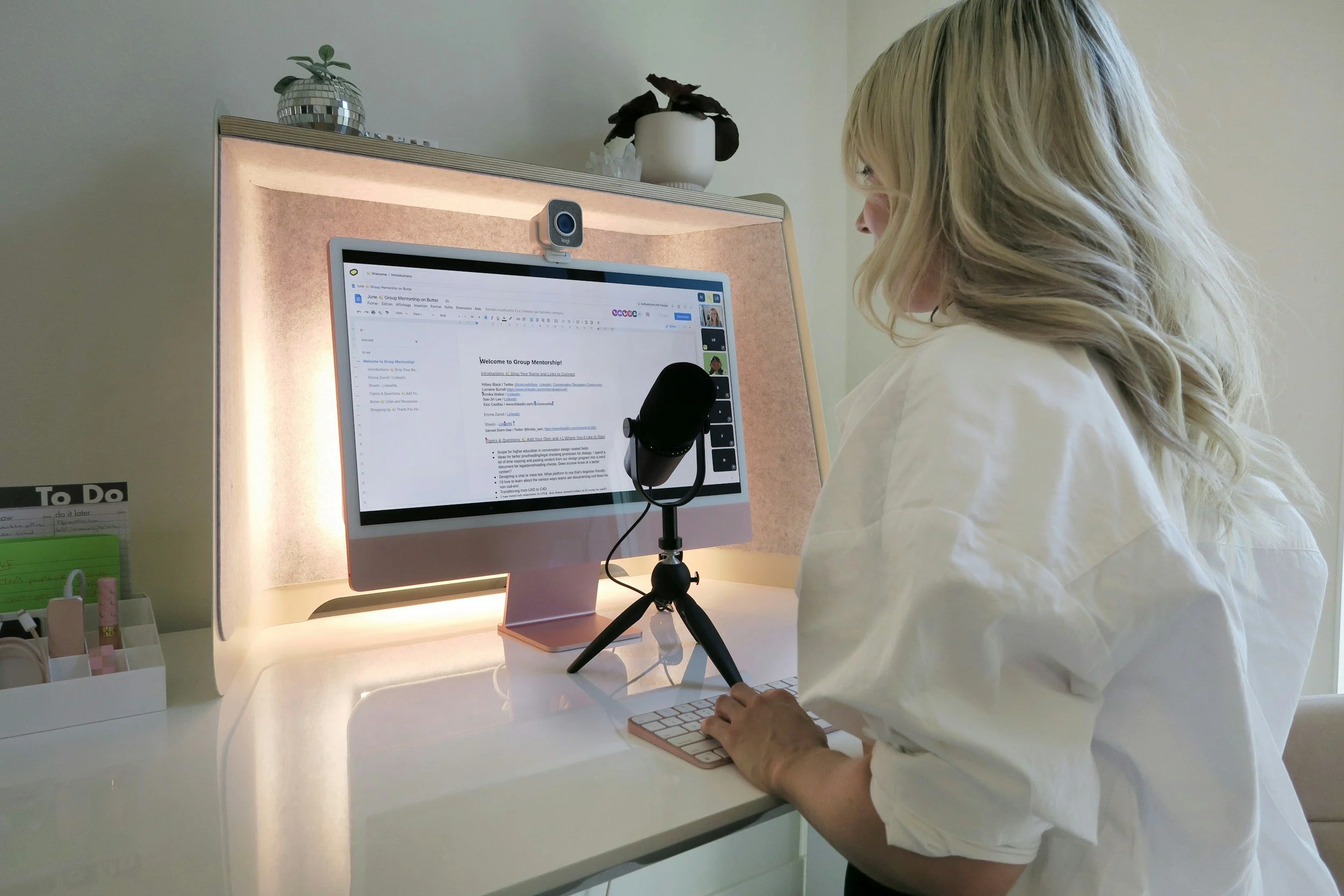 A person with blonde hair is seated at a white desk, using a computer with a large monitor. The monitor displays a document with text and a sidebar, and a webcam is positioned above the screen. A microphone on a small tripod and a wireless keyboard are also on the desk. The background features a pinkish panel, a potted plant, and a decorative object.