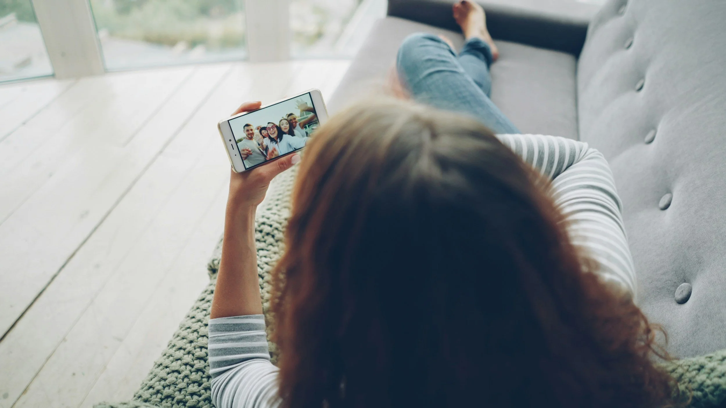 A woman with auburn hair lying on a gray sofa, holding a smartphone in her right hand, taking a selfie with a group of friends visible on the phone's screen. The sofa is located near a large glass window with a scenic outdoor view.