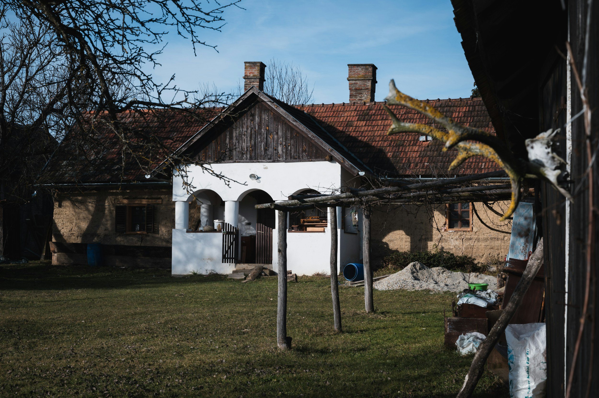 A rustic house with a red tiled roof, white and beige walls, and a small porch supported by white columns, situated in a grassy yard with leafless trees and a sunny sky.